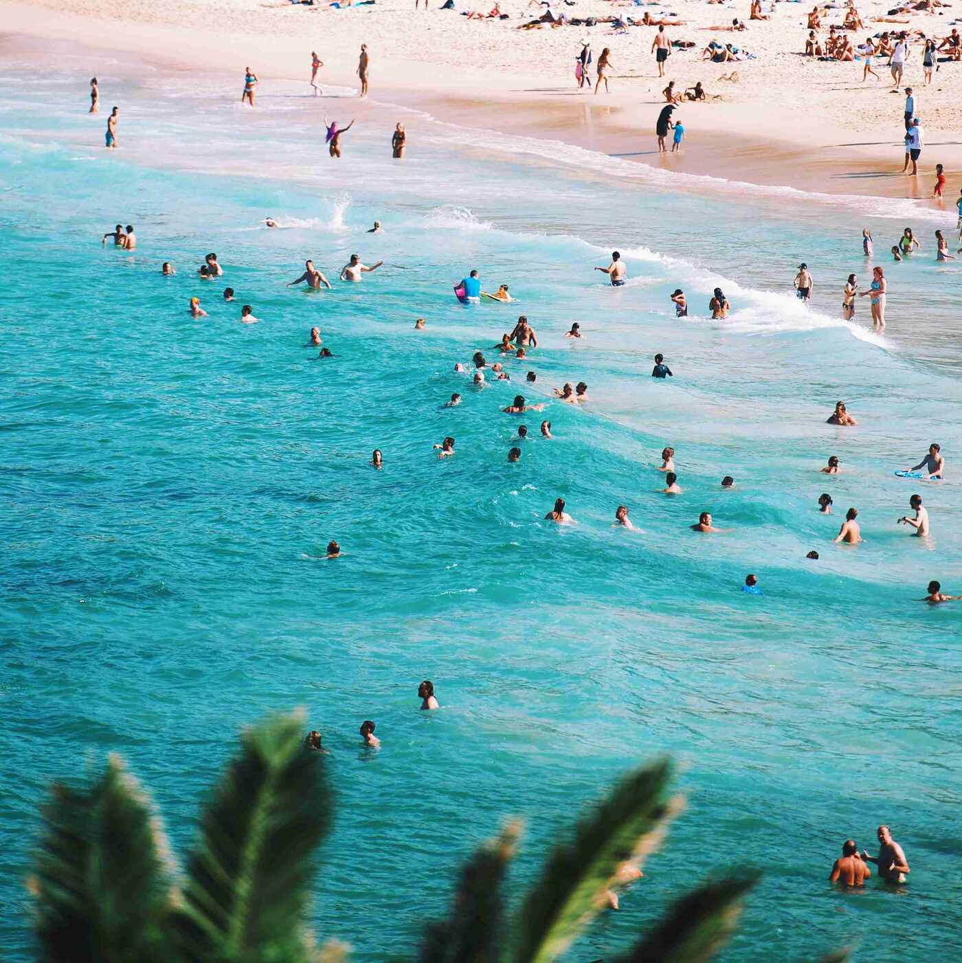Menschen schwimmen und spielen im klaren, türkisfarbenen Wasser eines Strandes, während andere am Strand entspannen. Im Hintergrund sind Sonnenanbeter auf dem Sand zu sehen, umgeben von üppigem Pflanzenbewuchs.