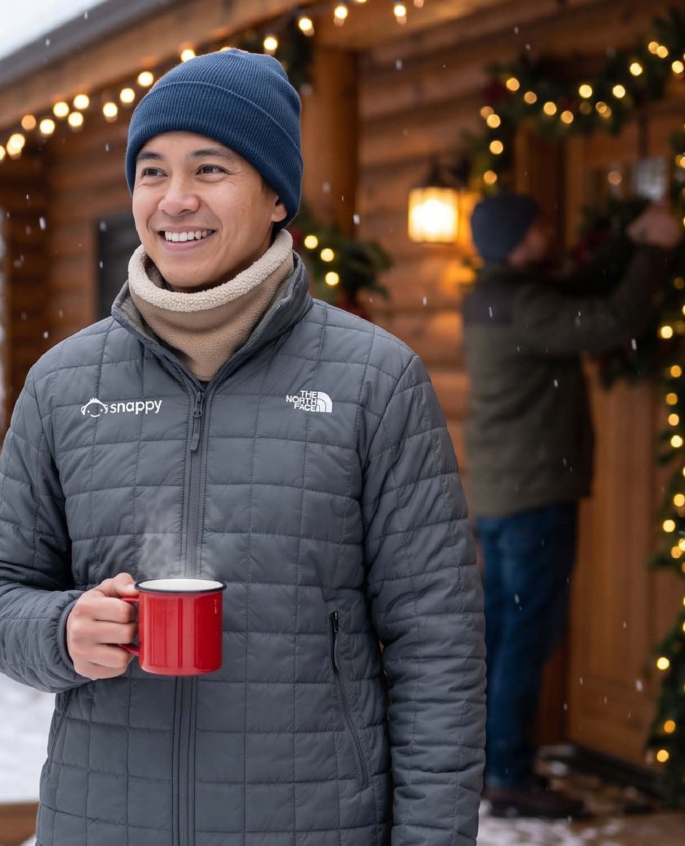 Smiling person in a Snappy-branded jacket holding a red mug outside a decorated cabin