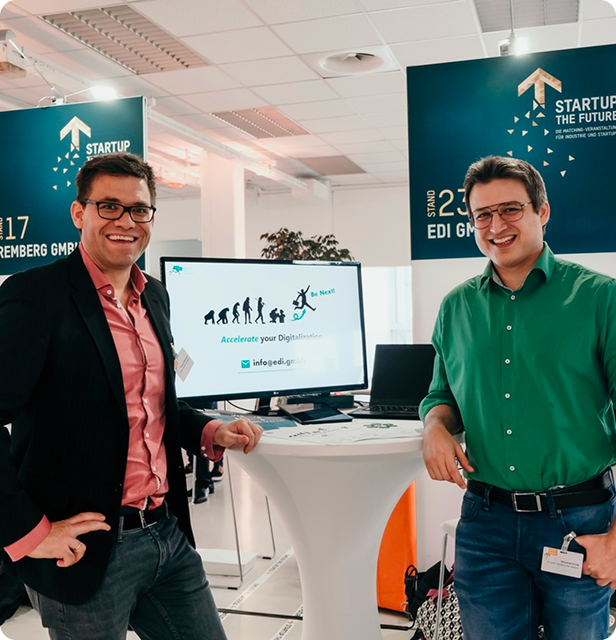 Thomas Freudenmann (left) and Mohanad El-Haji (right), co-founders of EDI GmbH, stand smiling at an event booth with a monitor displaying 'Accelerate your digital future' and a 'STARTUP THE FUTURE' banner in the background.
