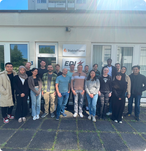 A diverse group of professionals from EDI GmbH stands outdoors in front of a modern building, smiling at the camera. The group includes individuals of various genders, ages, and appearances, reflecting the company's commitment to a global and diverse team.