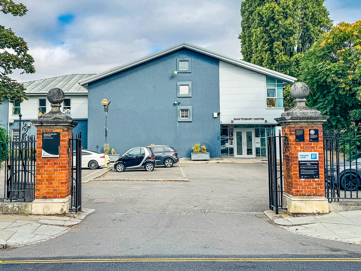 Entrance to Shaftesbury Centre with two brick pillars, black iron gates, and a parking lot with three cars in front of a blue and white building.