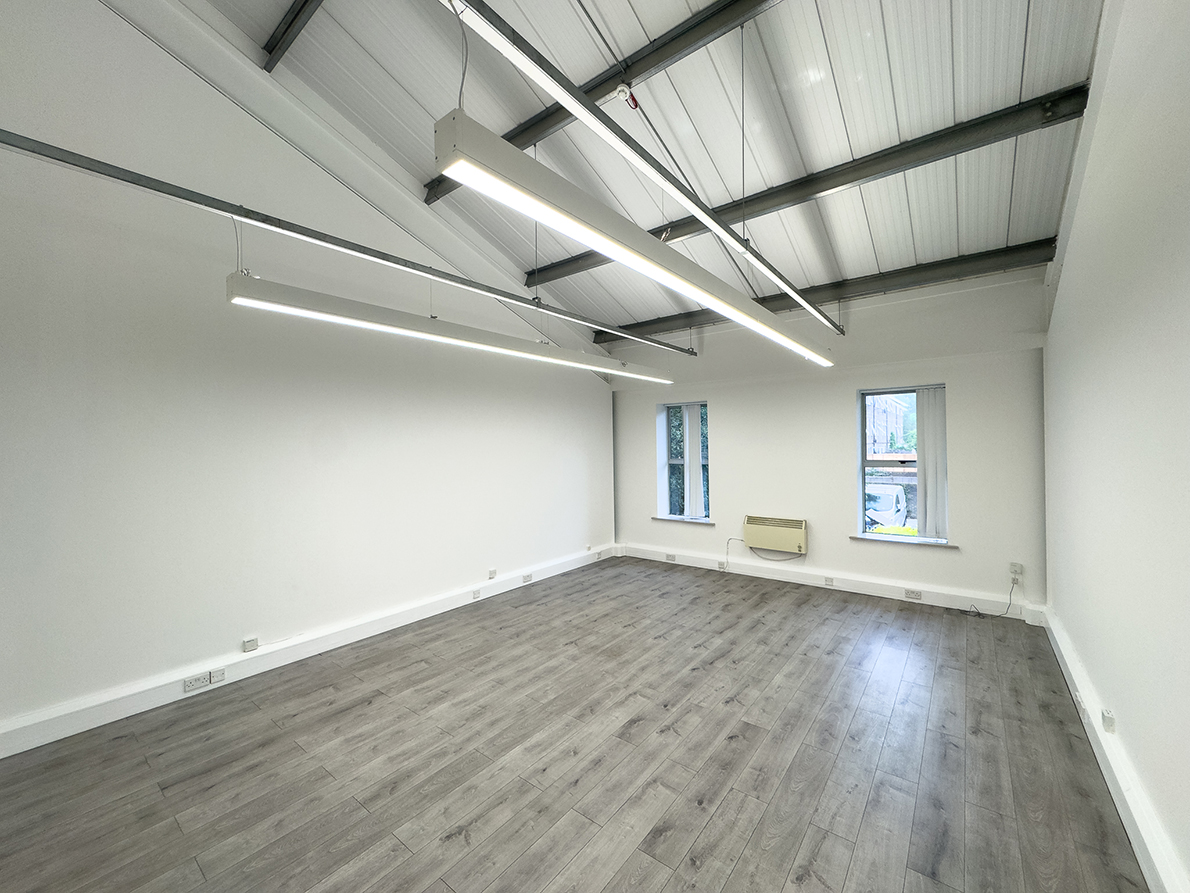 Empty room with light gray wood flooring, white walls, exposed ceiling beams, hanging fluorescent lights, and two windows on one wall.