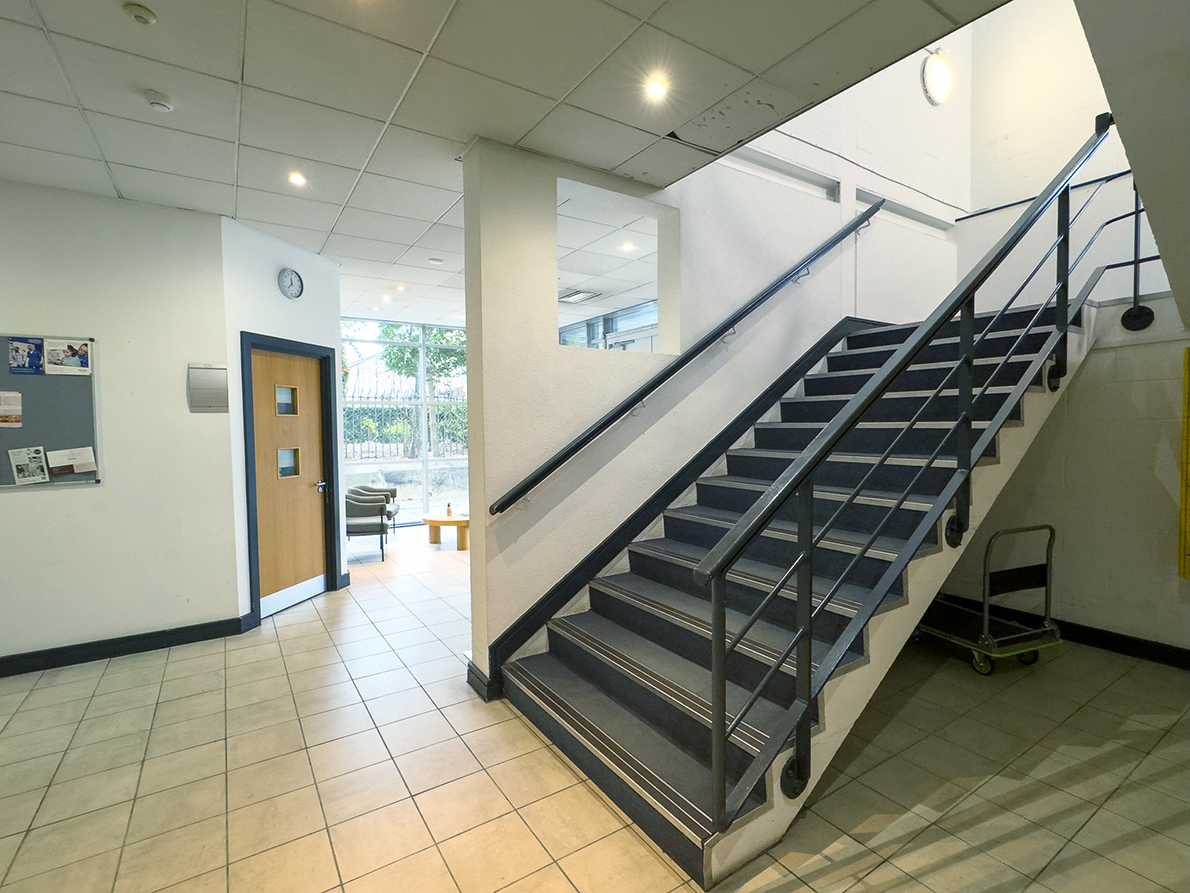 Indoor staircase with black metal railing next to a hallway leading to a room with chairs and a window.
