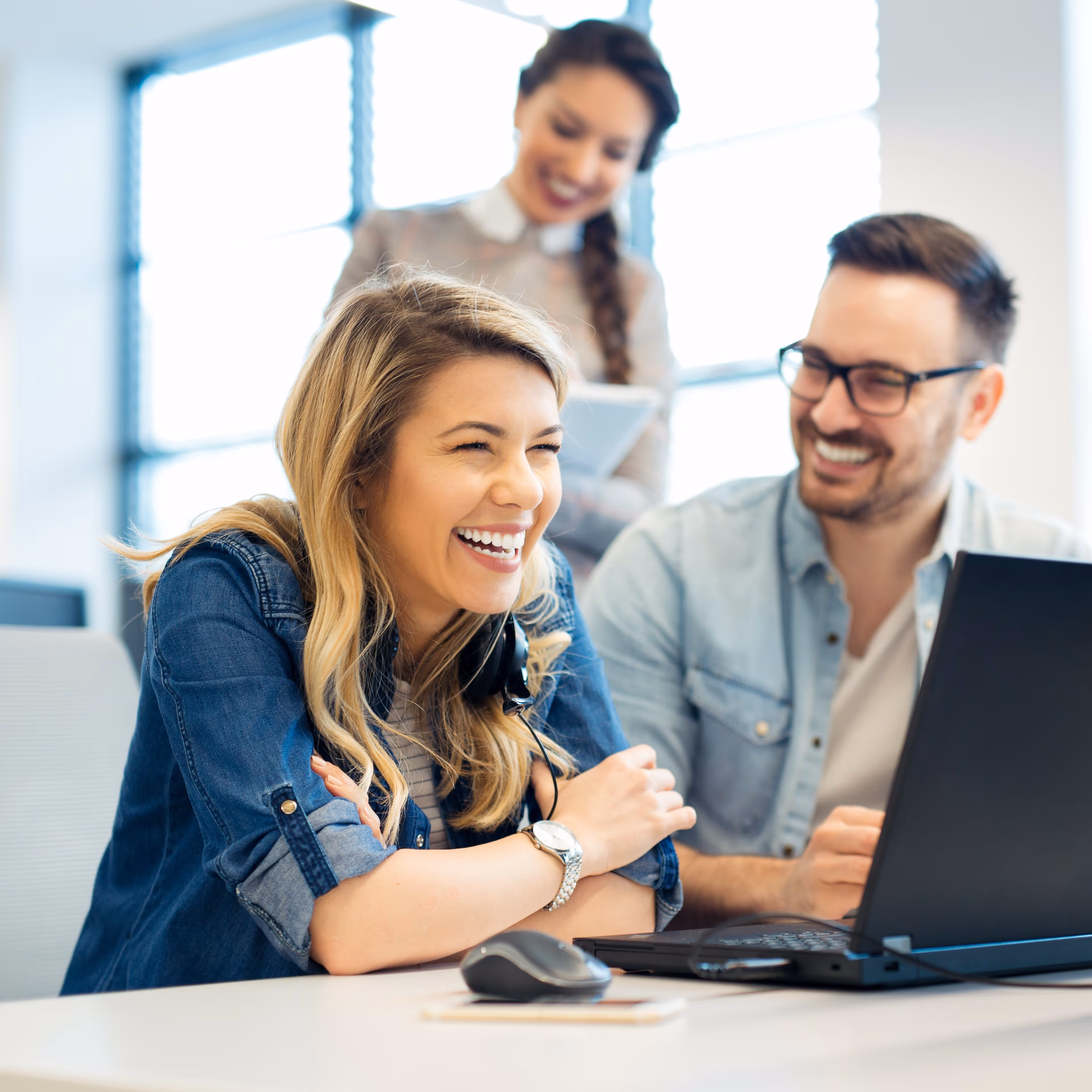 A trio of workers laugh as they work in an office, sharing a computer.