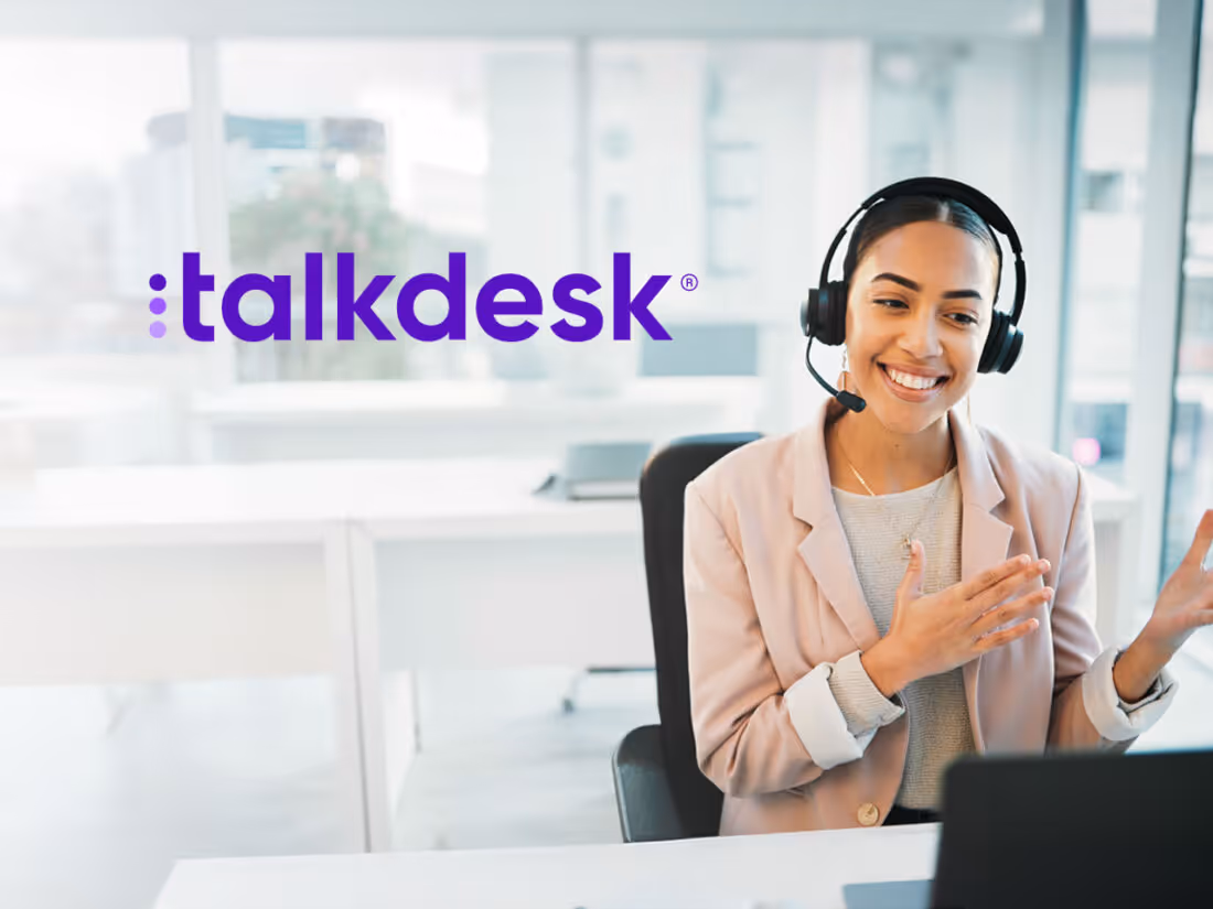 Photo of woman wearing a phone headset working at a laptop with the Talkdesk logo in the upper corner of the photo