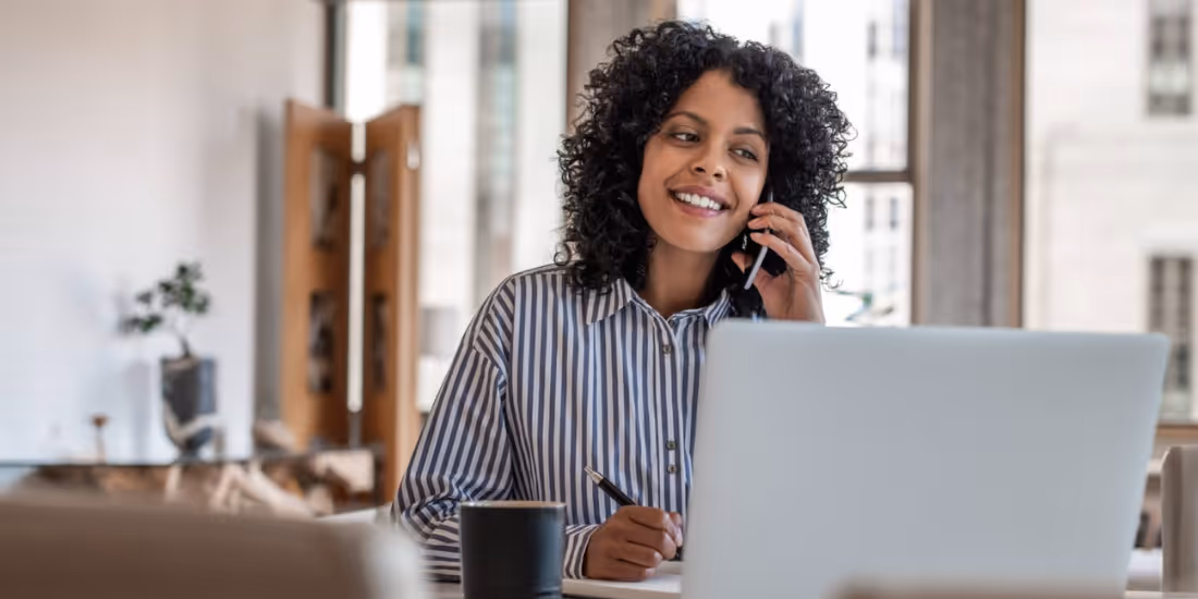 A woman sits in front of a computer with a pen while talking on a cell phone.