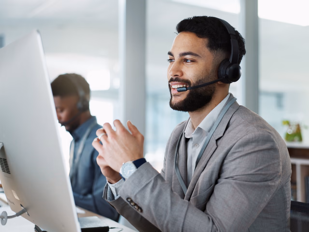 Smiling call center agent wearing headset and gray blazer, speaking while sitting at a desk with computer monitor.