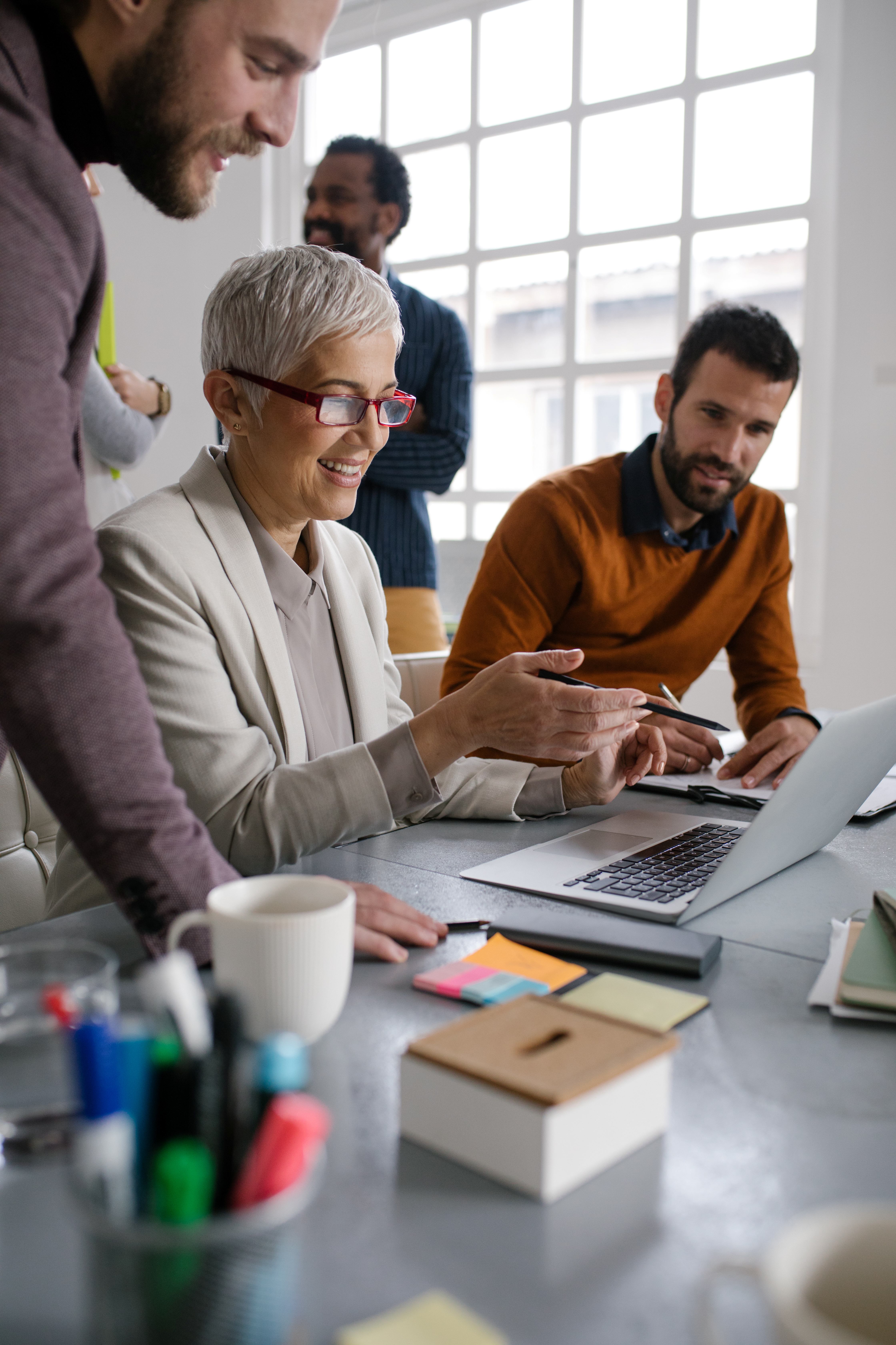 Three diverse coworkers discussing work around a laptop at a bright office table with stationery.
