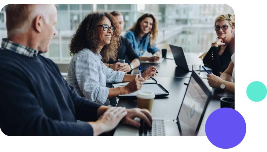 Team of four diverse coworkers smiling and collaborating at a desk with laptops and coffee cups.