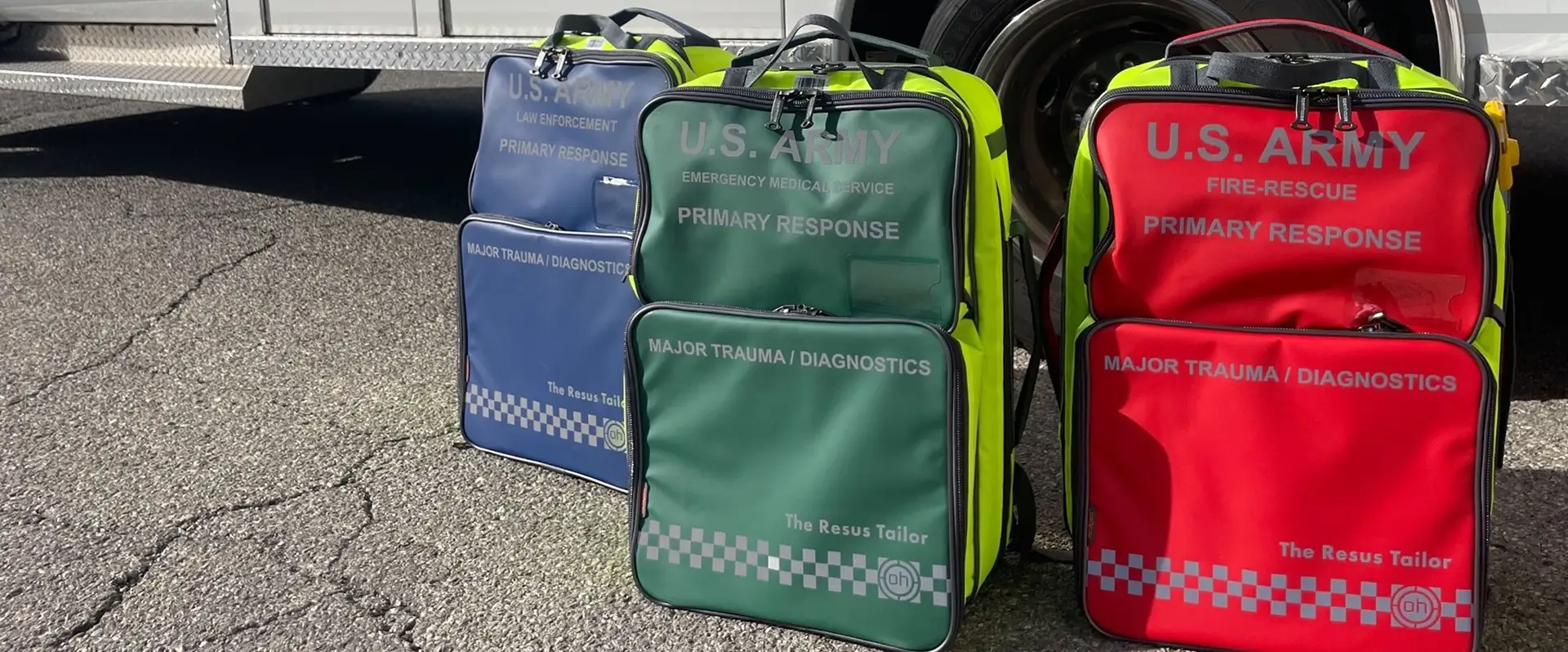 Three U.S. Army primary response medical bags in blue, green, and red for law enforcement, emergency medical service, and fire-rescue, arranged on asphalt.