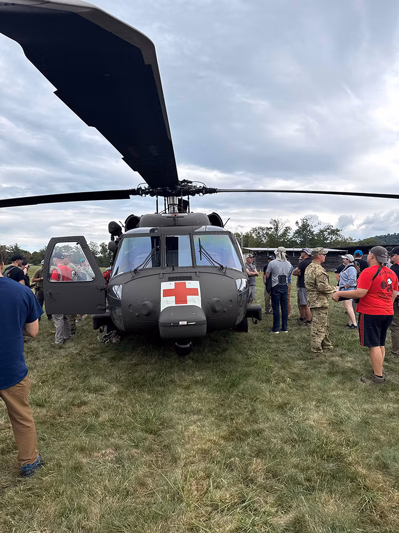 Military medical helicopter on grass with open door, surrounded by people, and cloudy sky.