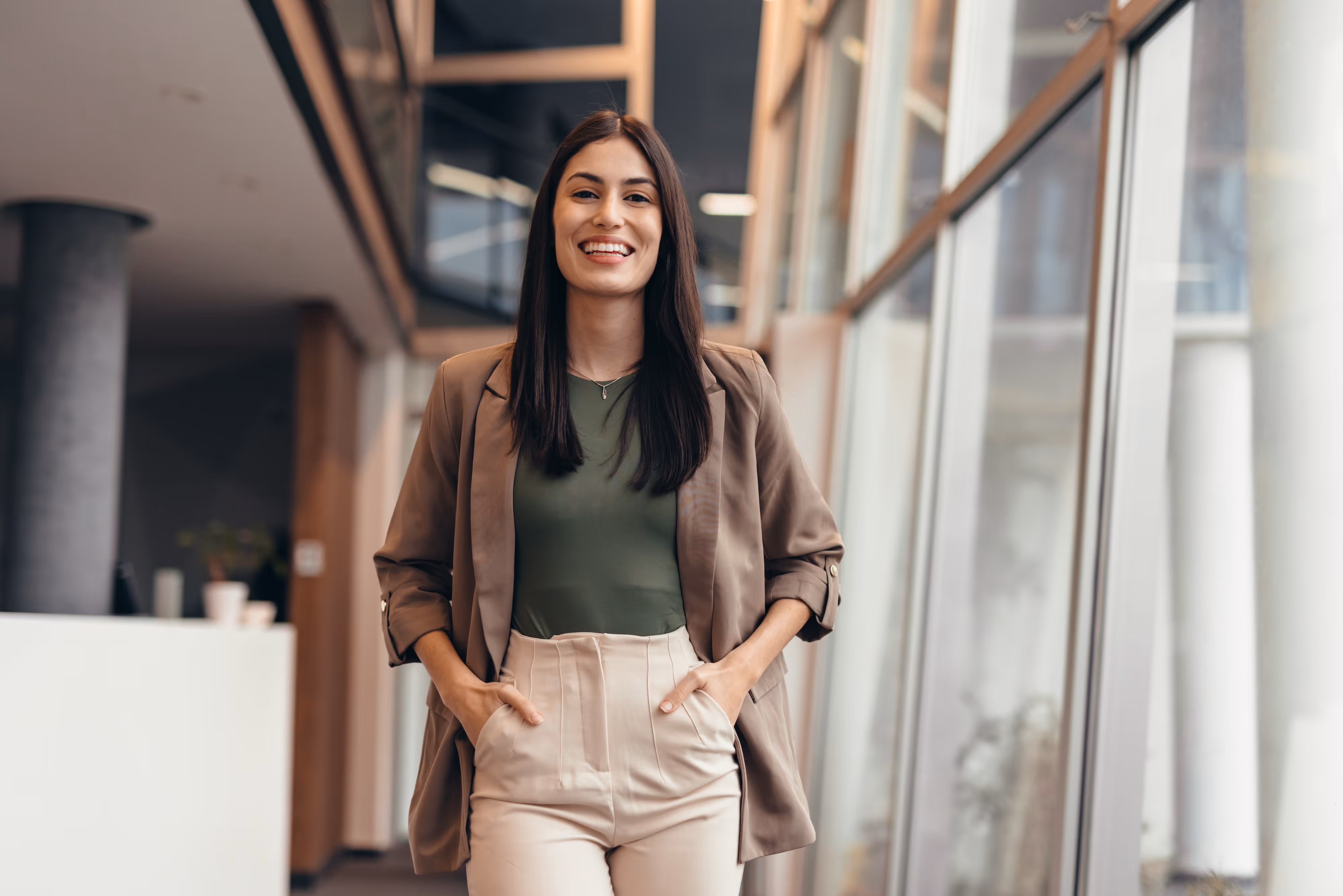 Smiling woman in a brown blazer and light pants standing in a modern office corridor with glass walls.