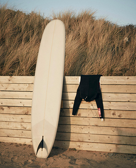 Planche de surf blanche appuyée contre une palissade en bois avec une combinaison noire suspendue à côté, sur une plage avec des herbes hautes en arrière-plan.