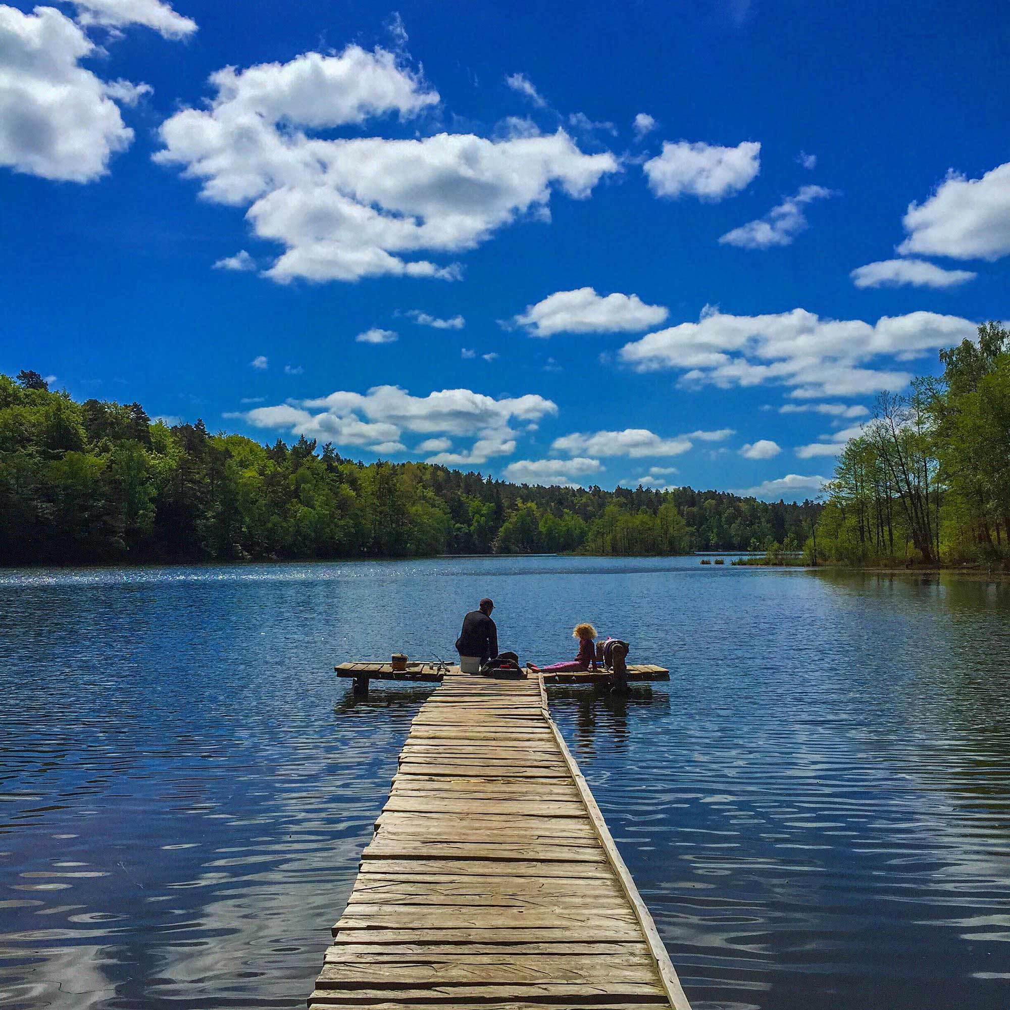 Zwei Personen sitzen am Ende eines Holzpontons auf einem See, umgeben von Bäumen unter blauem Himmel 