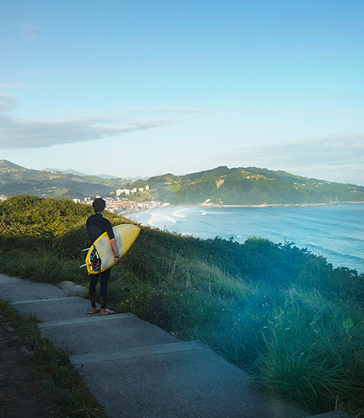 Surfeur tenant sa planche jaune regardant l'océan depuis un sentier surélevé au Pays Basque.