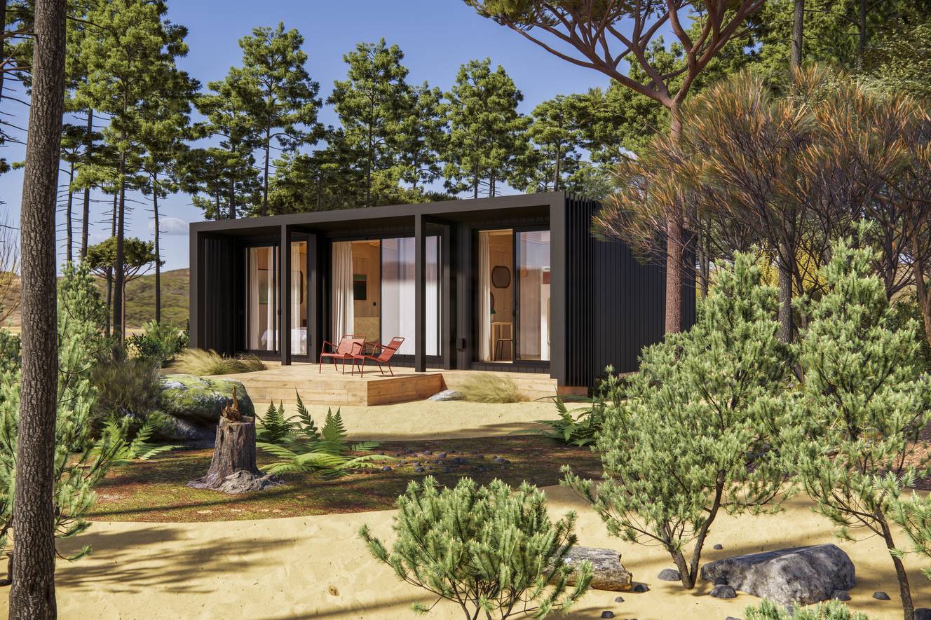 Petite villa moderne noire avec terrasse en bois et fauteuils rouges, entourée de pins et de buissons dans un paysage sablonneux sous un ciel bleu.