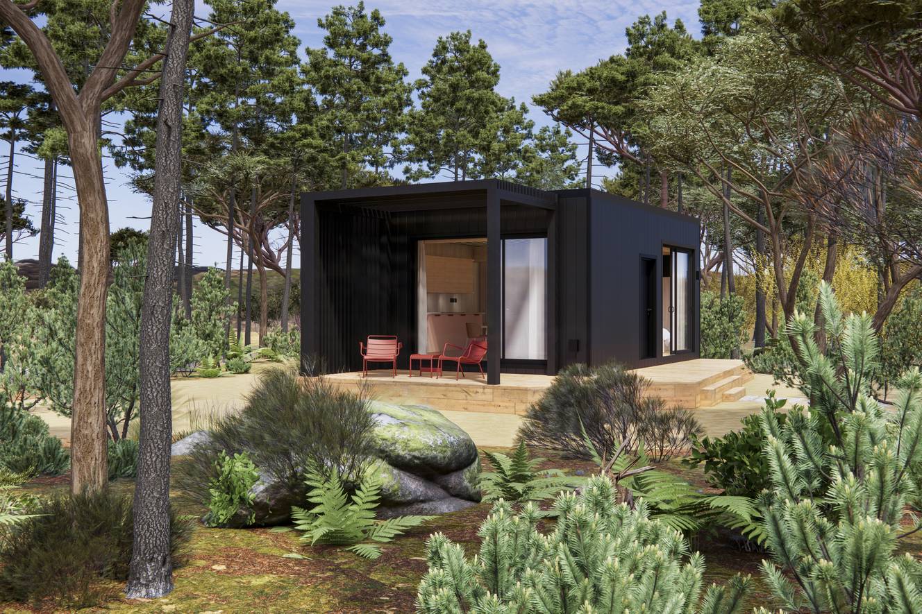 Petite maison moderne noire avec terrasse en bois entourée d’arbres et de végétation dense, avec deux chaises rouges sur la terrasse.