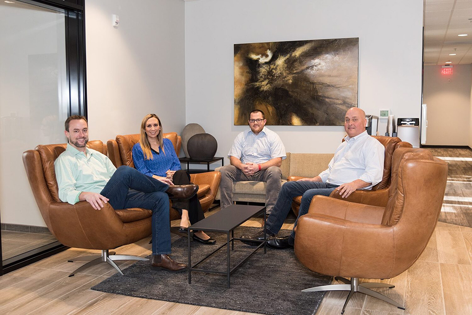 Four professionals are seated in a modern office lounge area. Two men are in brown leather armchairs, one wearing a light green shirt and the other in a white shirt. A woman in a blue blouse sits in the center, with a man in a light blue shirt seated on a beige couch to her right. A black coffee table is in front of them, and a large abstract painting hangs on the wall behind. The space is well-lit and features contemporary decor.