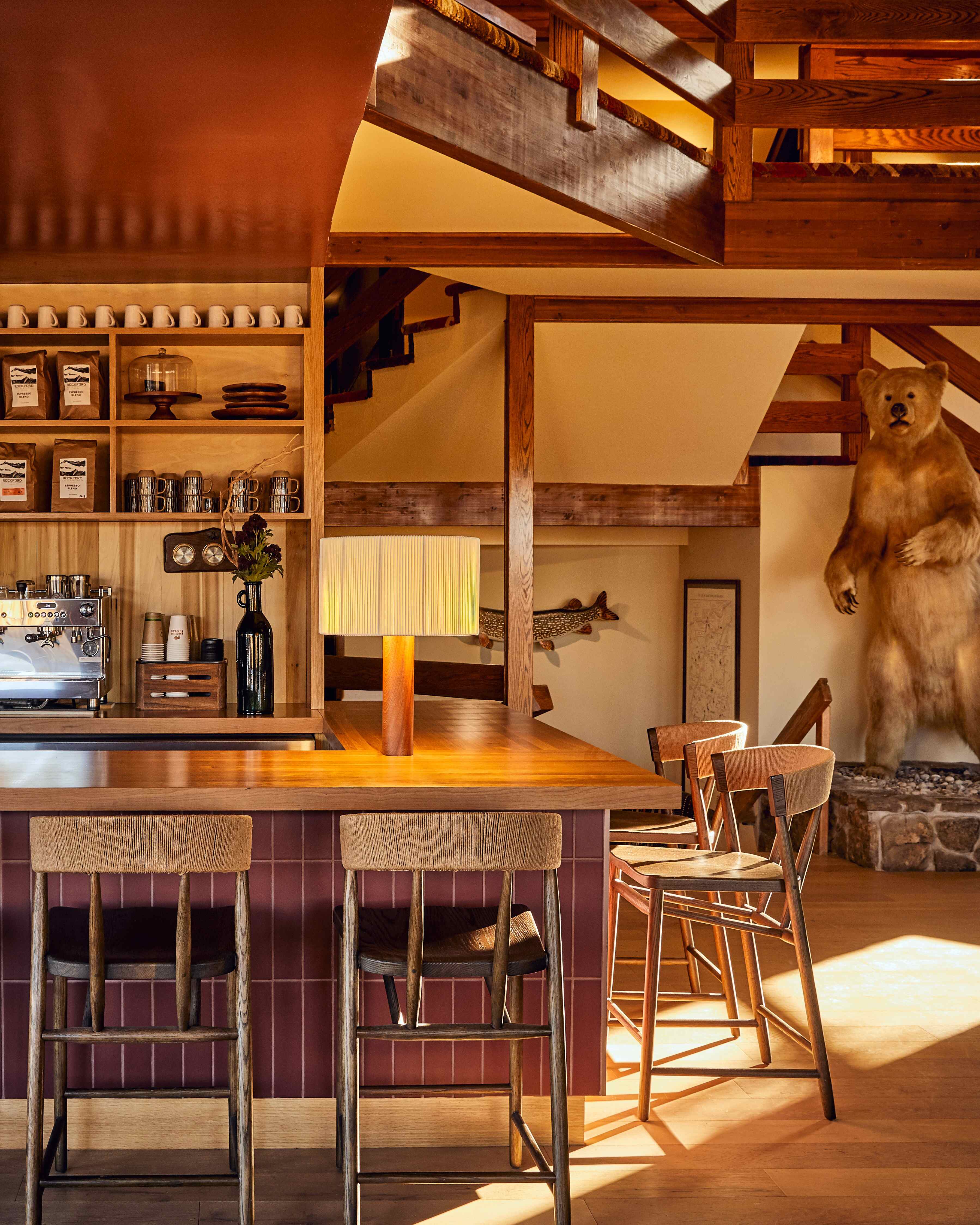 Modern coffee bar with wooden counter, high chairs, espresso machine, and shelves stocked with mugs and coffee bags.