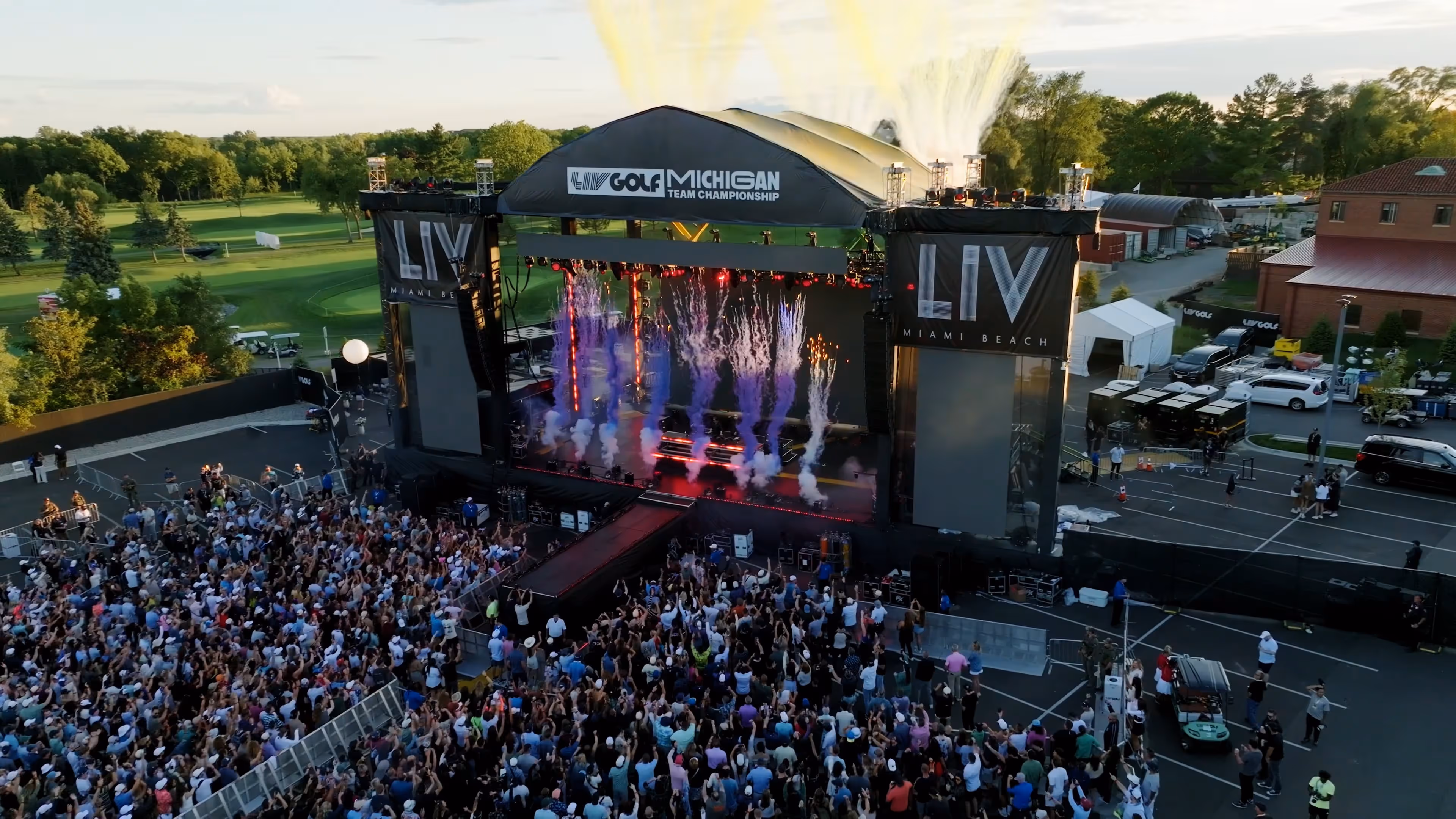 An aerial view captures the energy of a live concert staged at the LIV Golf Michigan Team Championship. The expansive outdoor stage is branded with bold signage for LIV Golf and LIV Miami Beach, framed by massive video screens and lighting rigs. Colorful pyrotechnics—purple smoke, orange sparks, and yellow bursts—shoot upward, creating a dramatic effect above the stage. A dense crowd of spectators fills the foreground, hands raised in excitement as the performance unfolds. Behind the stage, the green fairways of the golf course stretch into the distance, contrasting with the vibrant, high-energy atmosphere of the event.