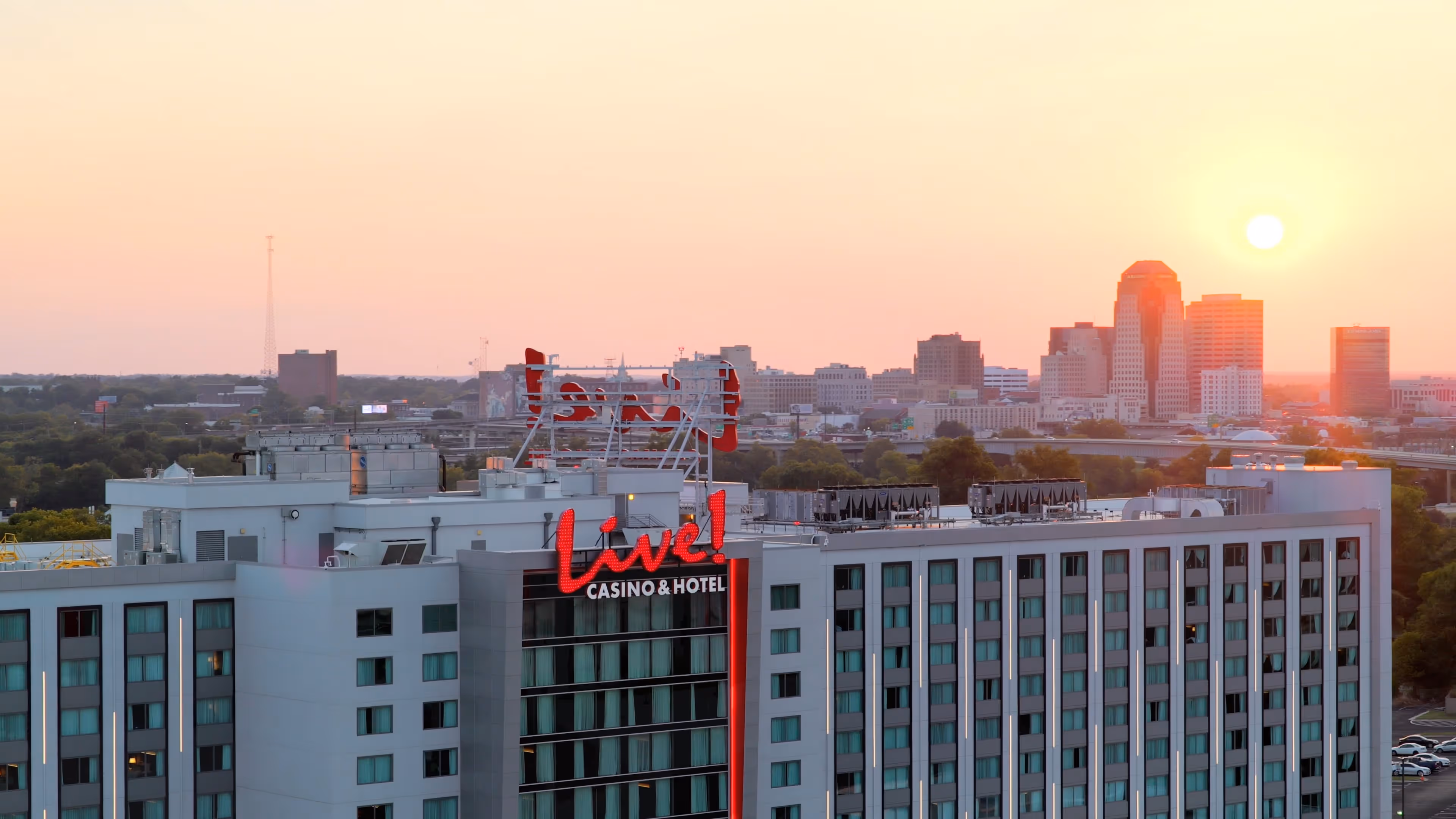 An elevated view captures the Live! Casino & Hotel in the foreground with its bold red signage illuminated against the soft evening light. The modern high-rise building features rows of reflective windows and a rooftop dotted with mechanical structures. Beyond the hotel, the city skyline rises with a cluster of mid- and high-rise buildings, their silhouettes glowing in the warm orange and pink hues of sunset. The sun hovers low on the horizon, casting a golden wash across the entire scene, blending the urban architecture with the natural gradient of the sky.