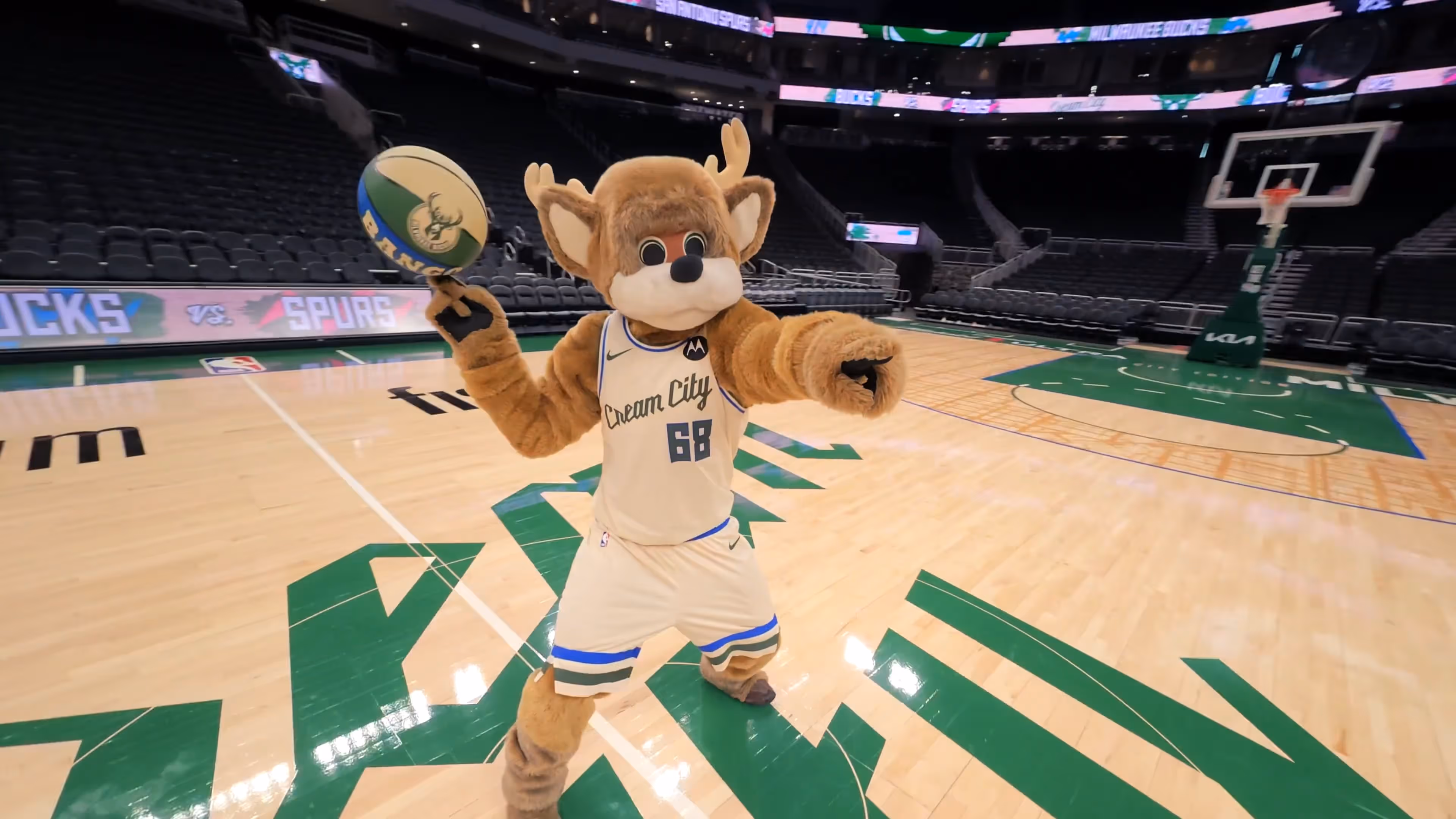 Basketball arena with mascot dressed as a deer wearing Cream City jersey number 68 spinning a basketball on finger.