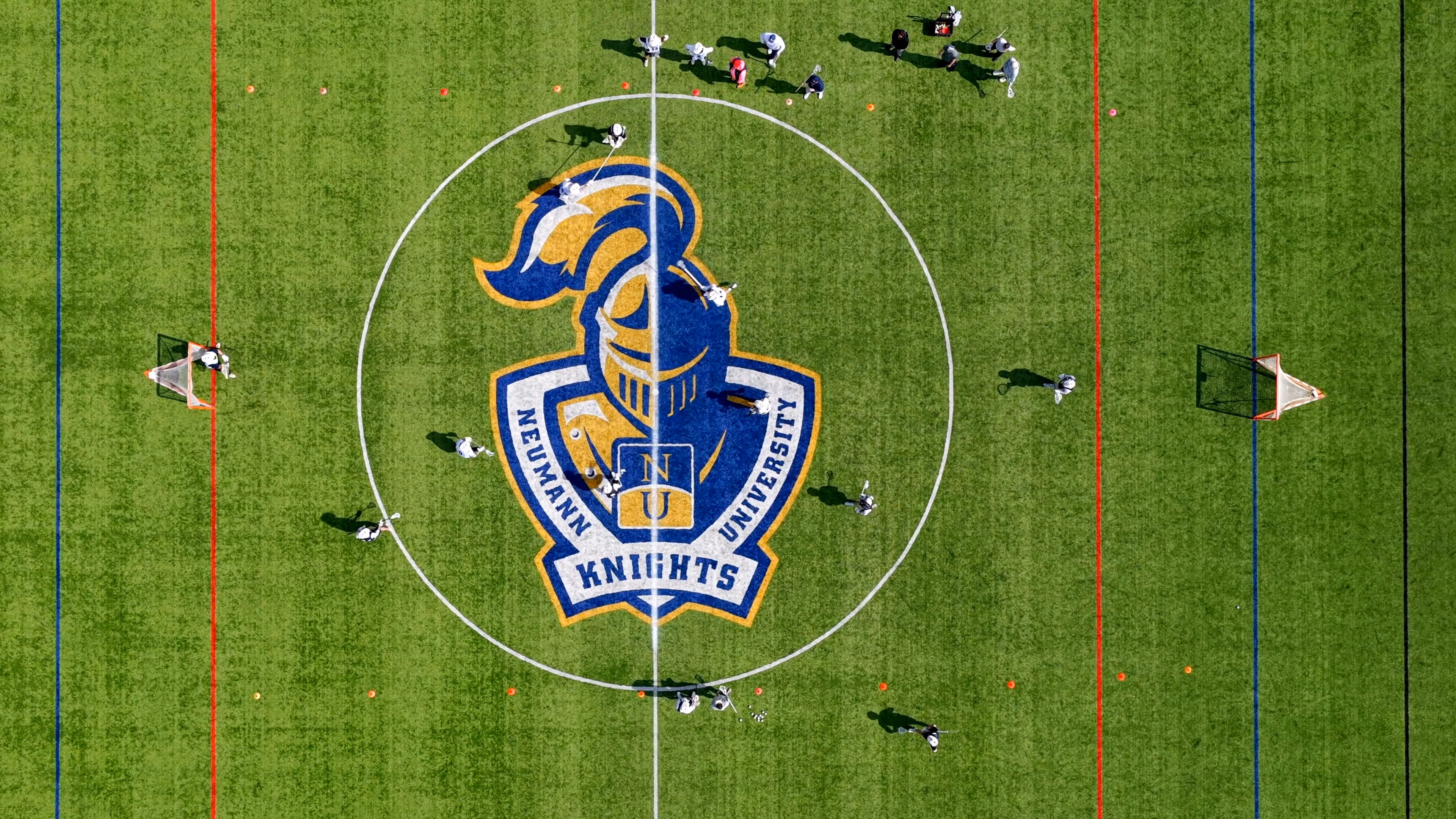 Aerial view of a lacrosse game in progress on a field with the Neumann University Knights logo at center.