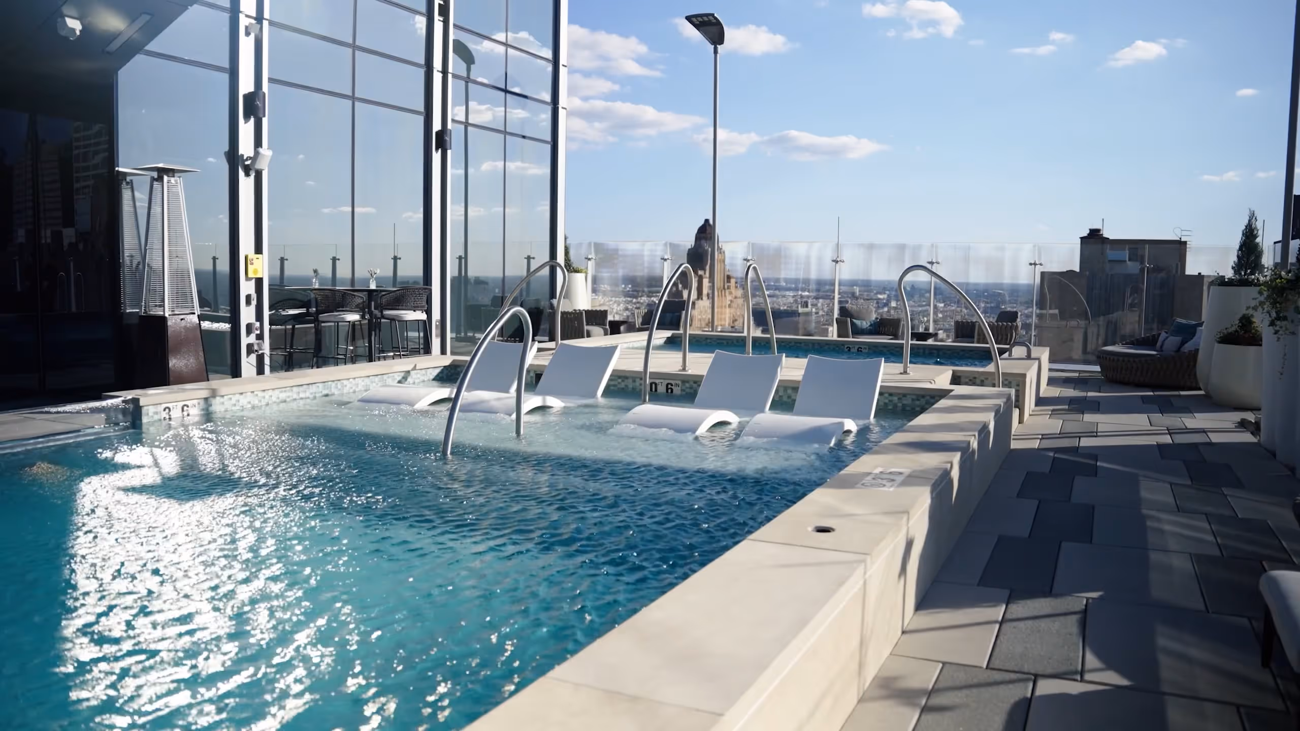 Rooftop pool area with submerged lounge chairs, glass railing, and city skyline in the background under clear blue sky.