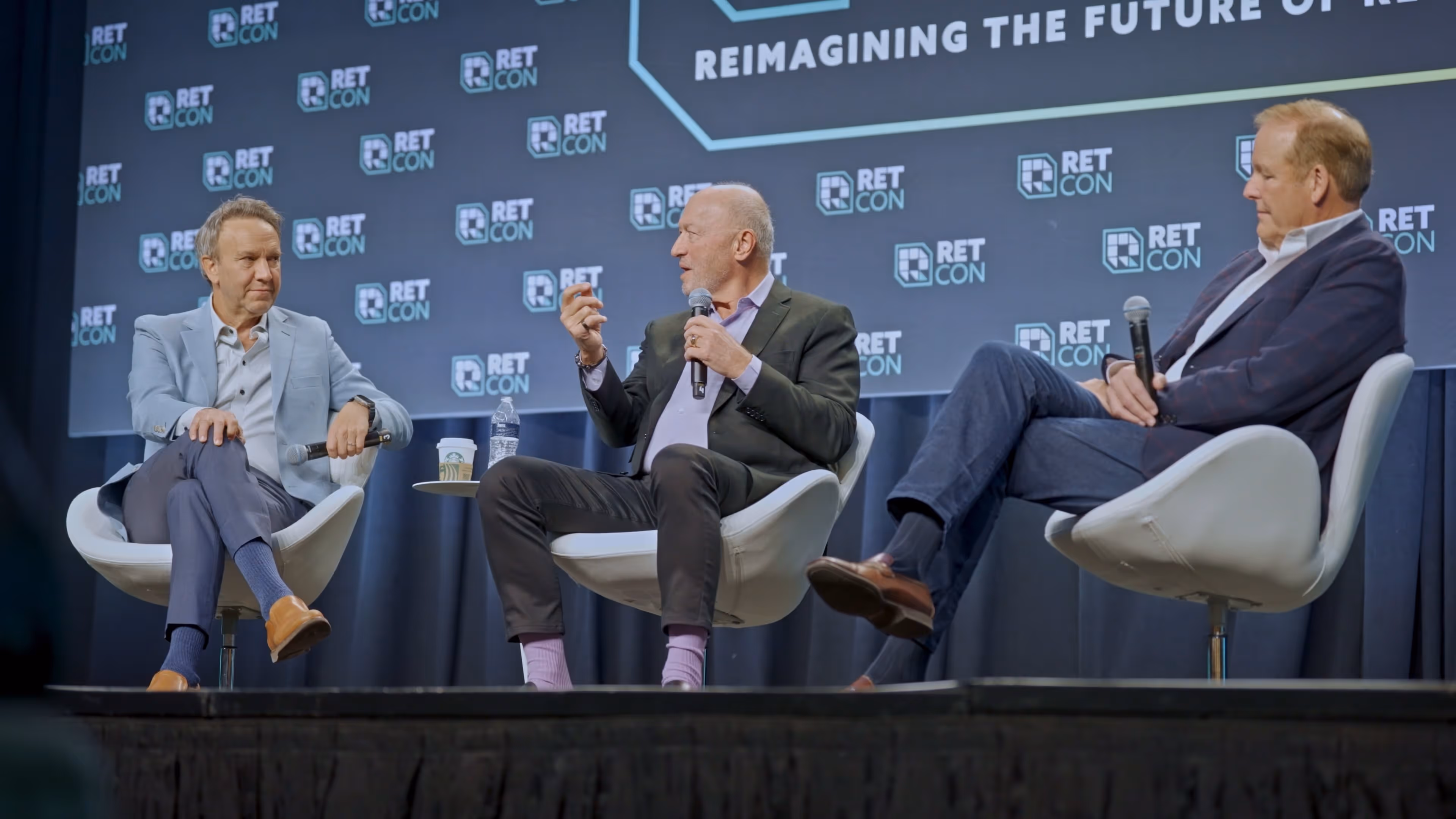 Three men in business attire seated on stage chairs during a panel discussion at RETCON event.