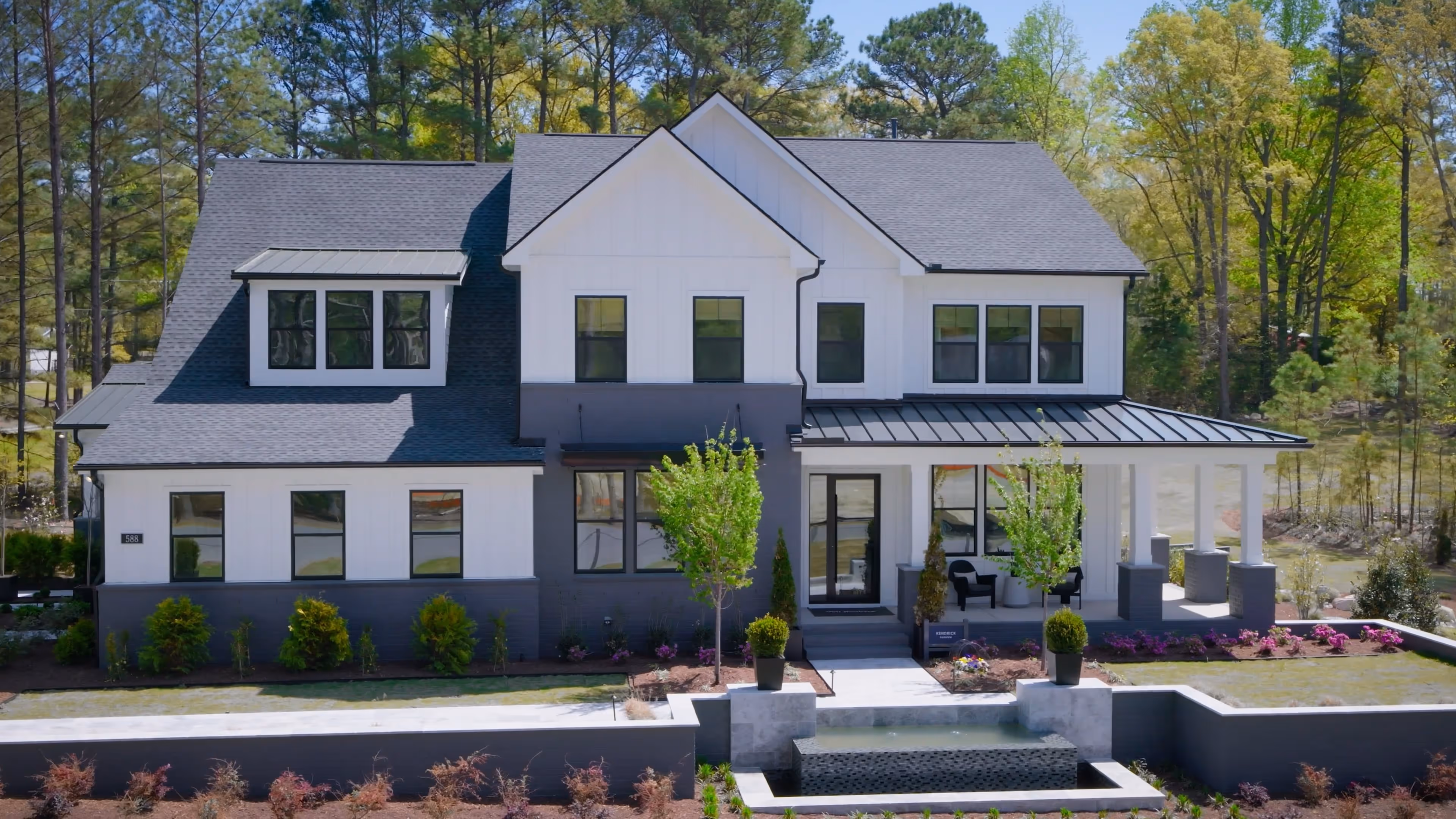 Modern two-story house with white and gray exterior, large windows, a porch with chairs, landscaped yard, and a water feature in front.