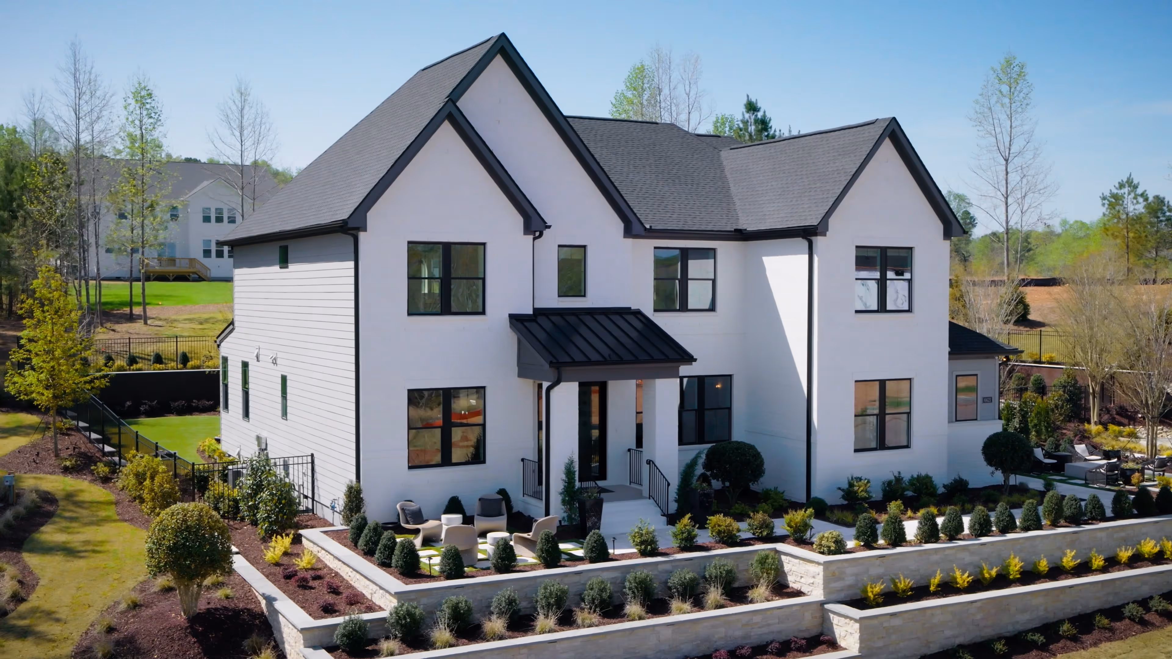 Modern two-story white house with black roof, black window frames, and a landscaped front yard with shrubs and outdoor seating.