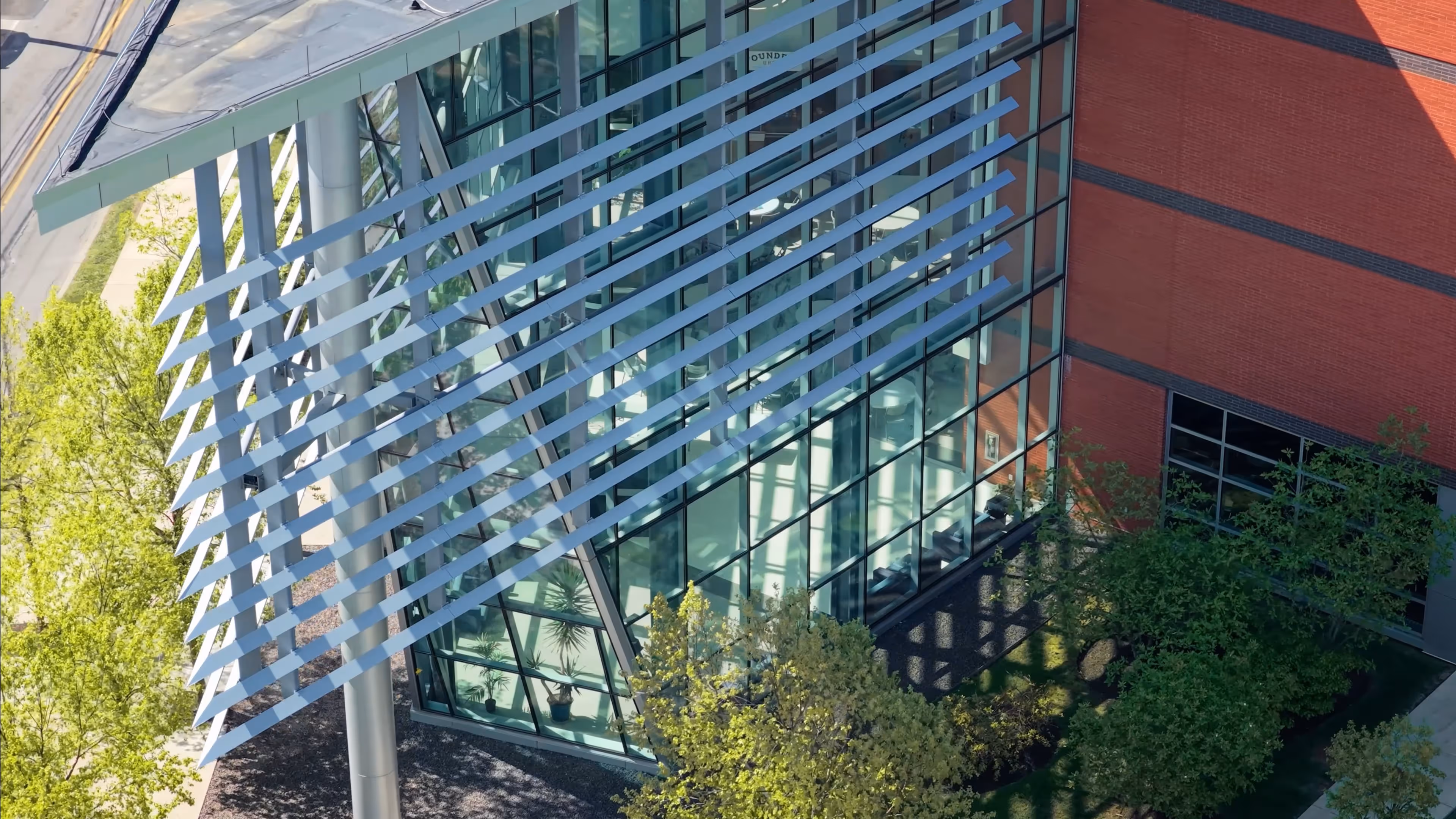 Modern glass building corner with angled metal sunshades and surrounding green trees.