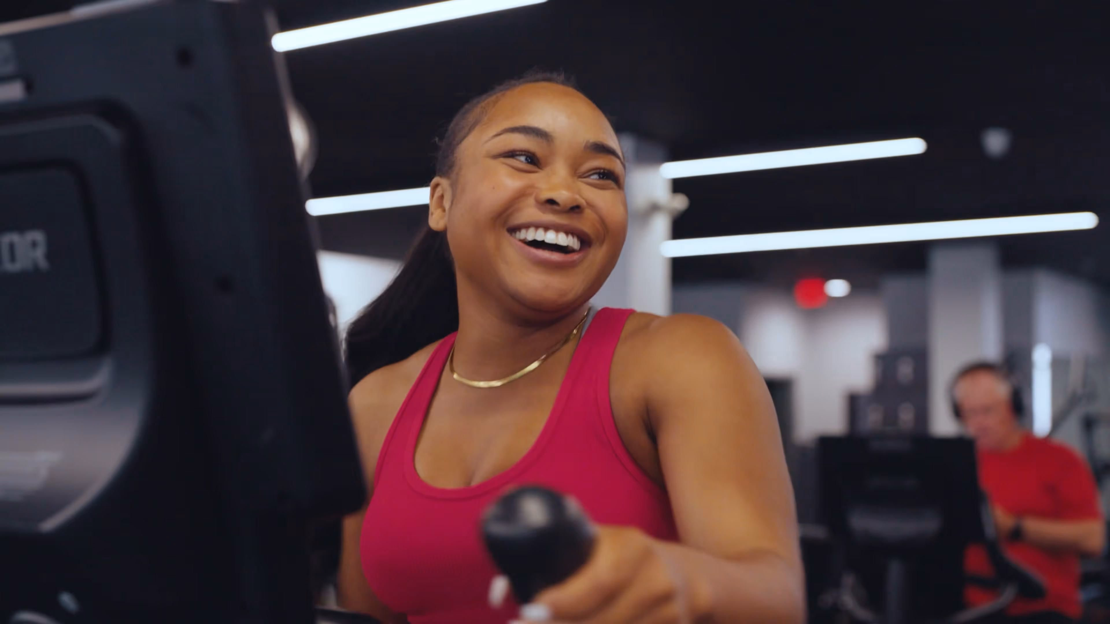 Smiling woman in a pink tank top exercising on a machine at the gym.