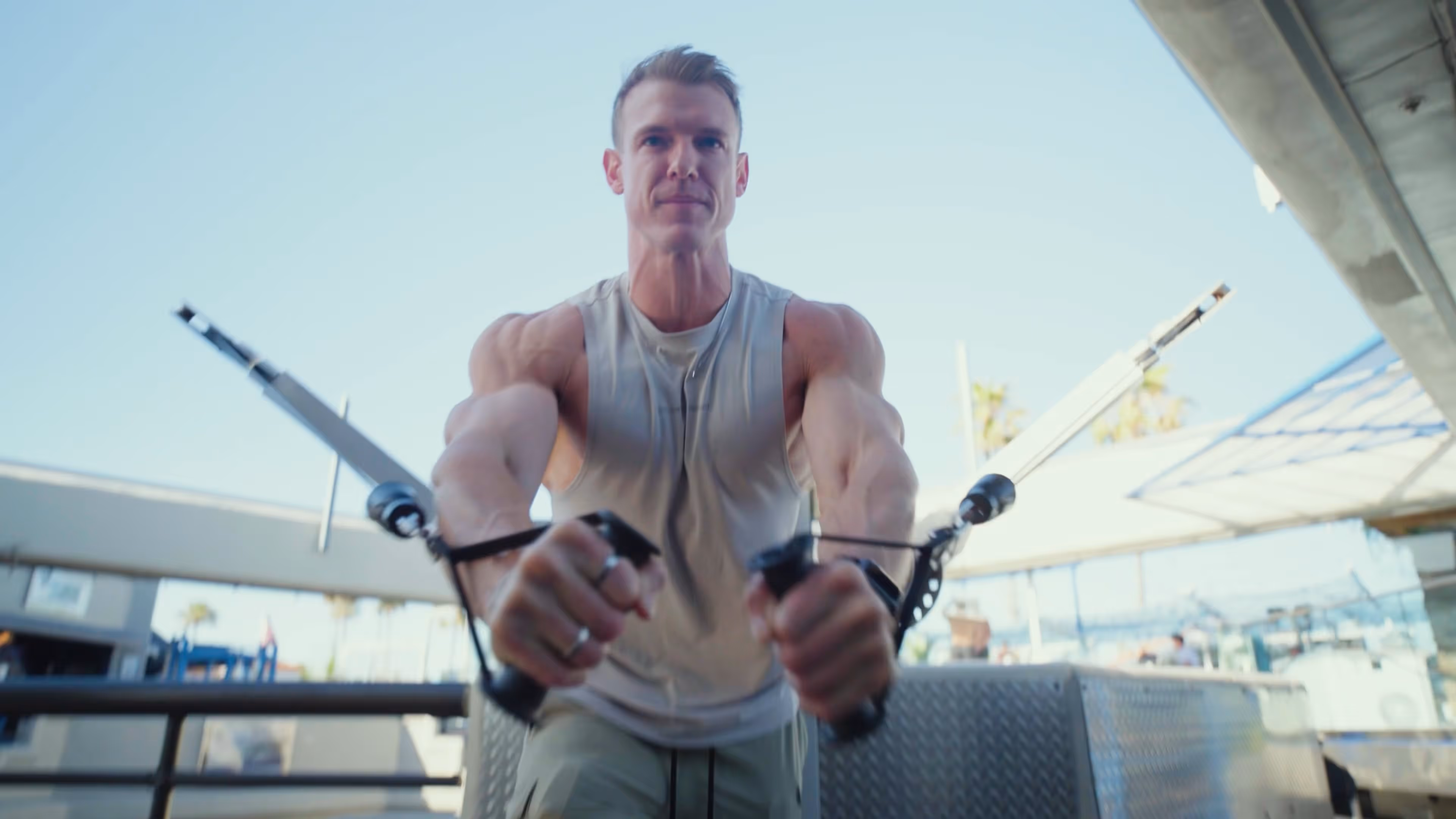 Muscular man in sleeveless shirt using cable machine for workout at outdoor gym.