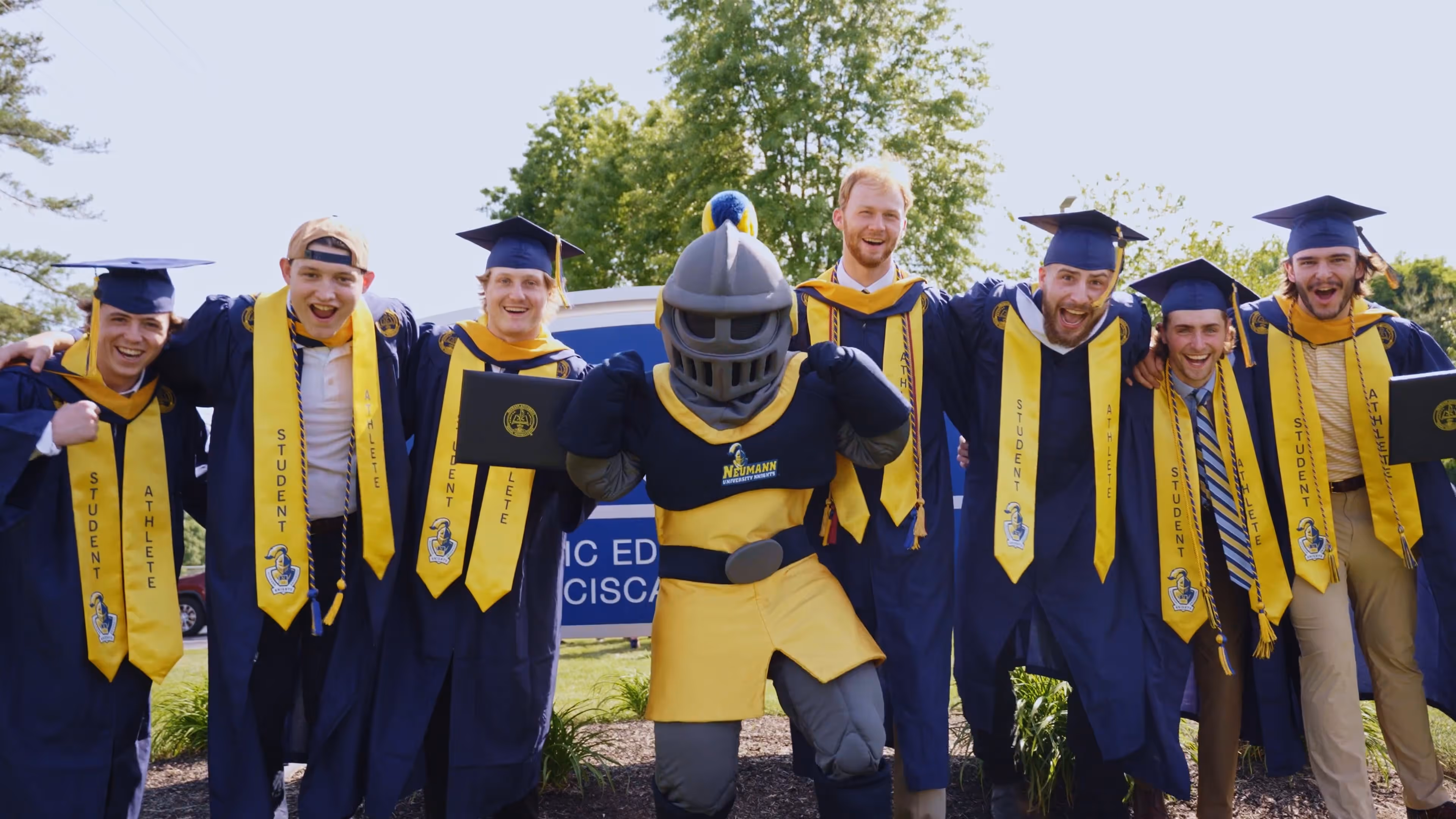 Group of male graduates in caps, gowns, and Student Athlete stoles posing with school mascot in daylight.
