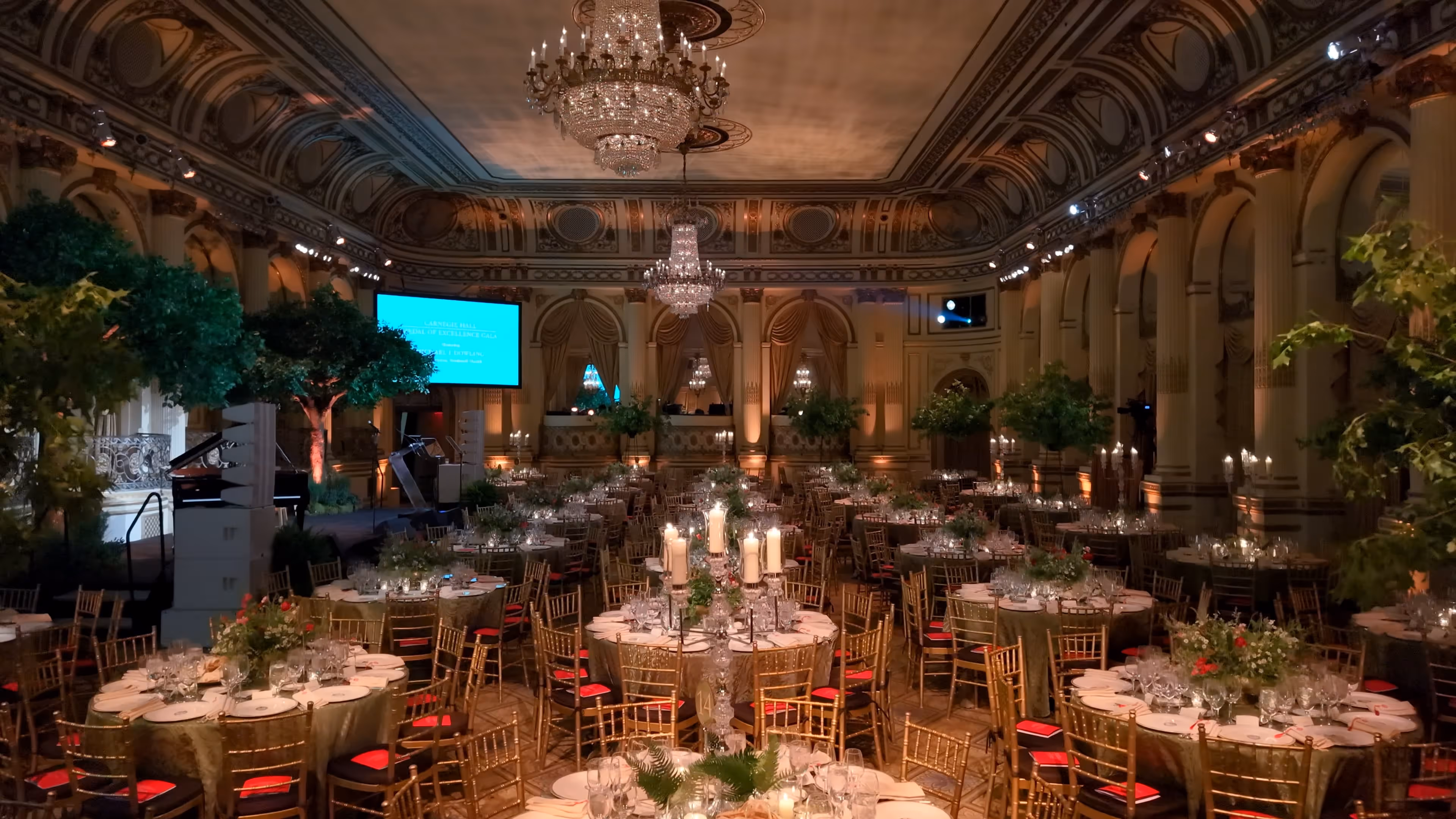Elegant ballroom banquet setup with round tables, gold chairs, chandeliers, candles, and floral centerpieces.