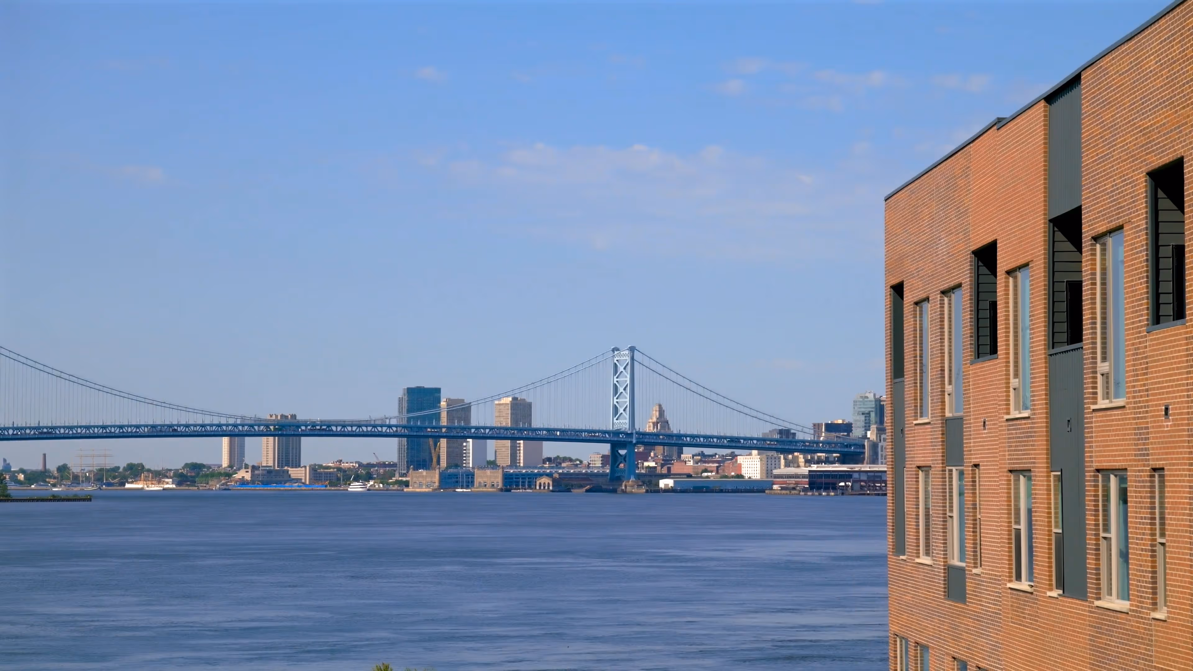 Blue suspension bridge over wide river with city skyline and brick building on the right.