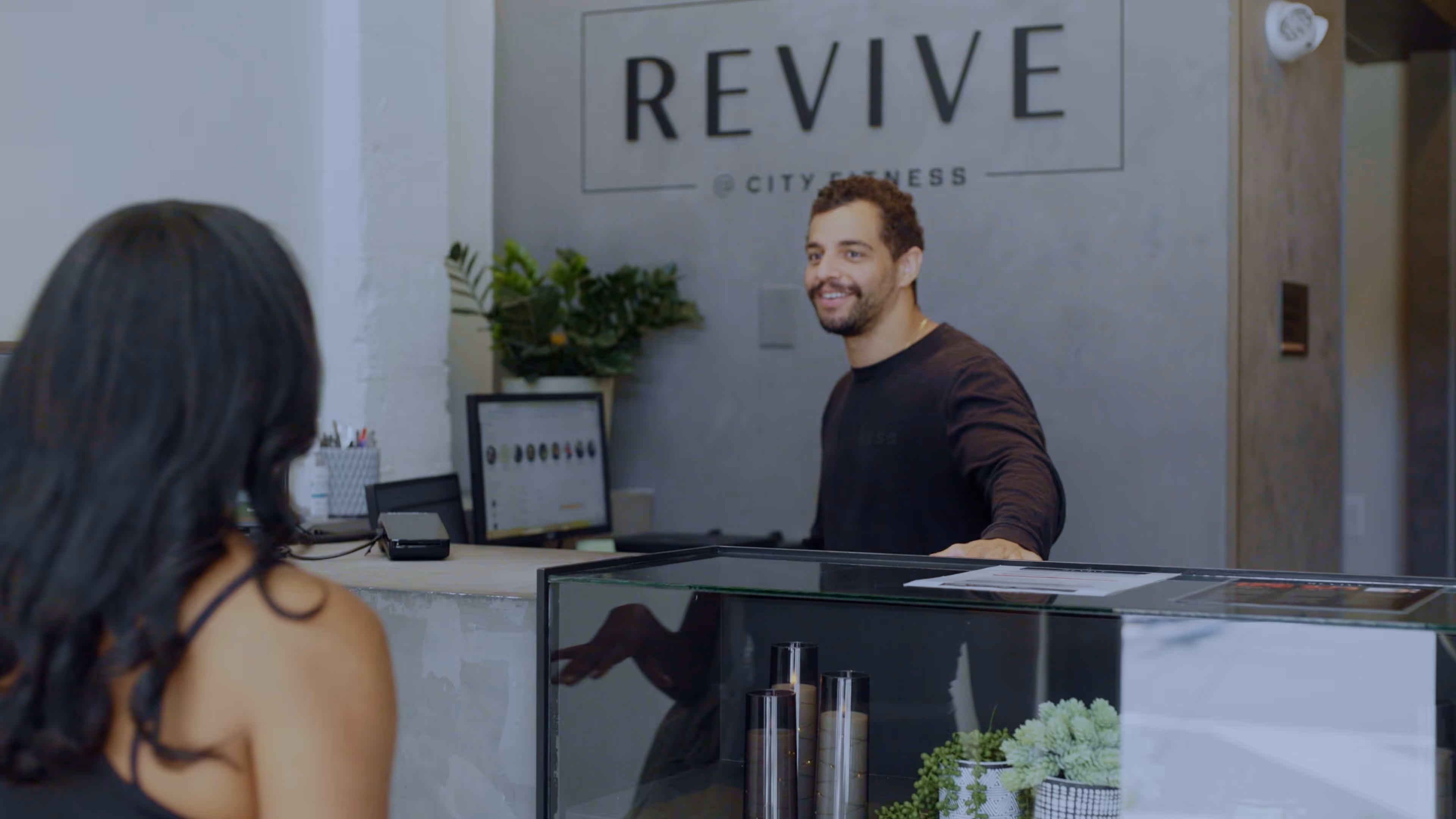 Man smiling behind reception desk at Revive City Fitness talking to woman in black top.