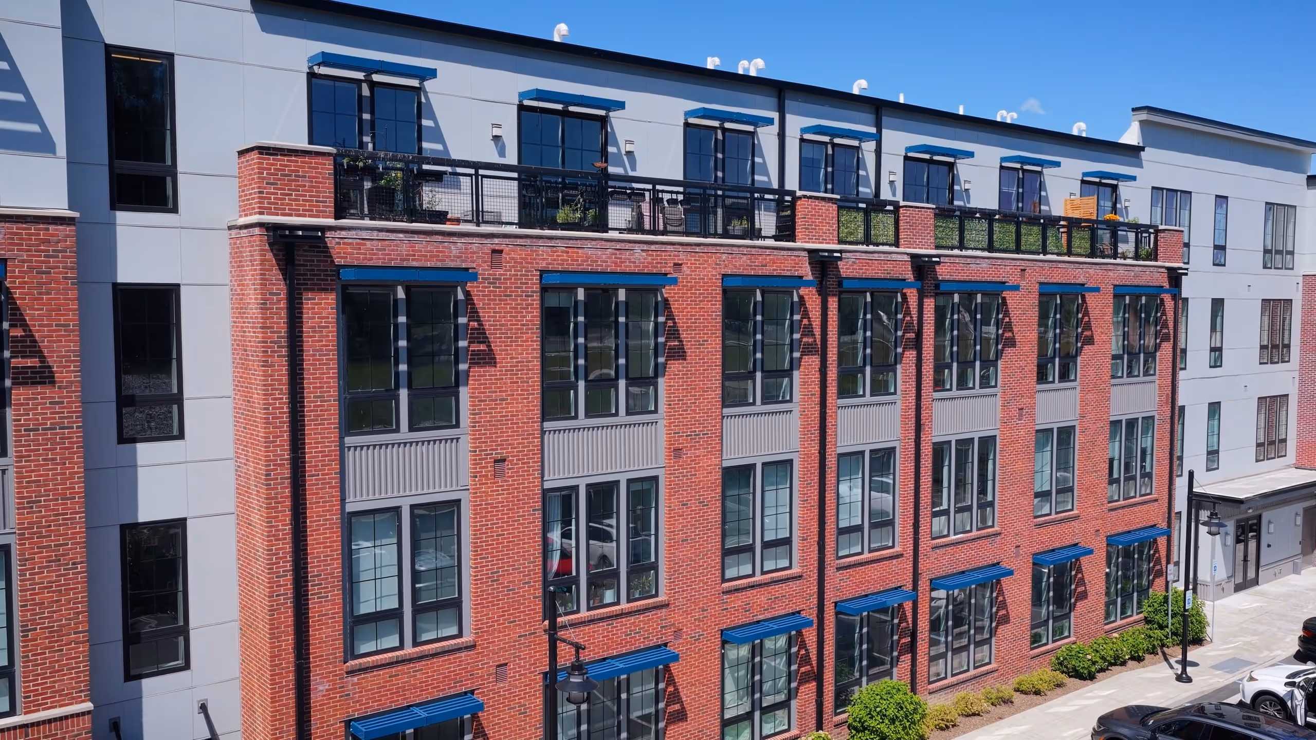 Modern red brick apartment building with black window frames, blue awnings, and rooftop balcony on sunny day.