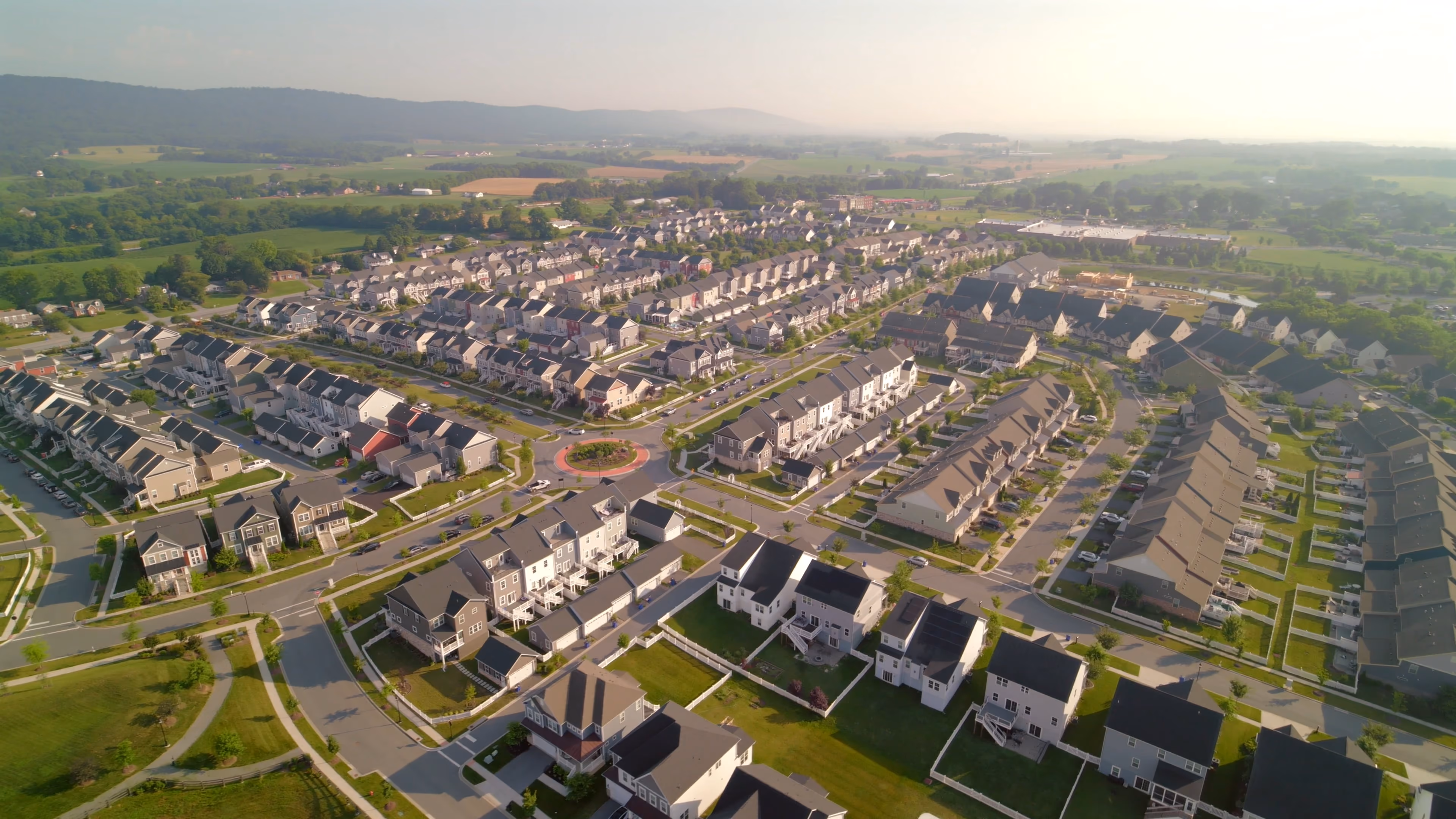 Aerial view of a suburban neighborhood with houses, streets, roundabout, and green fields in the distance.