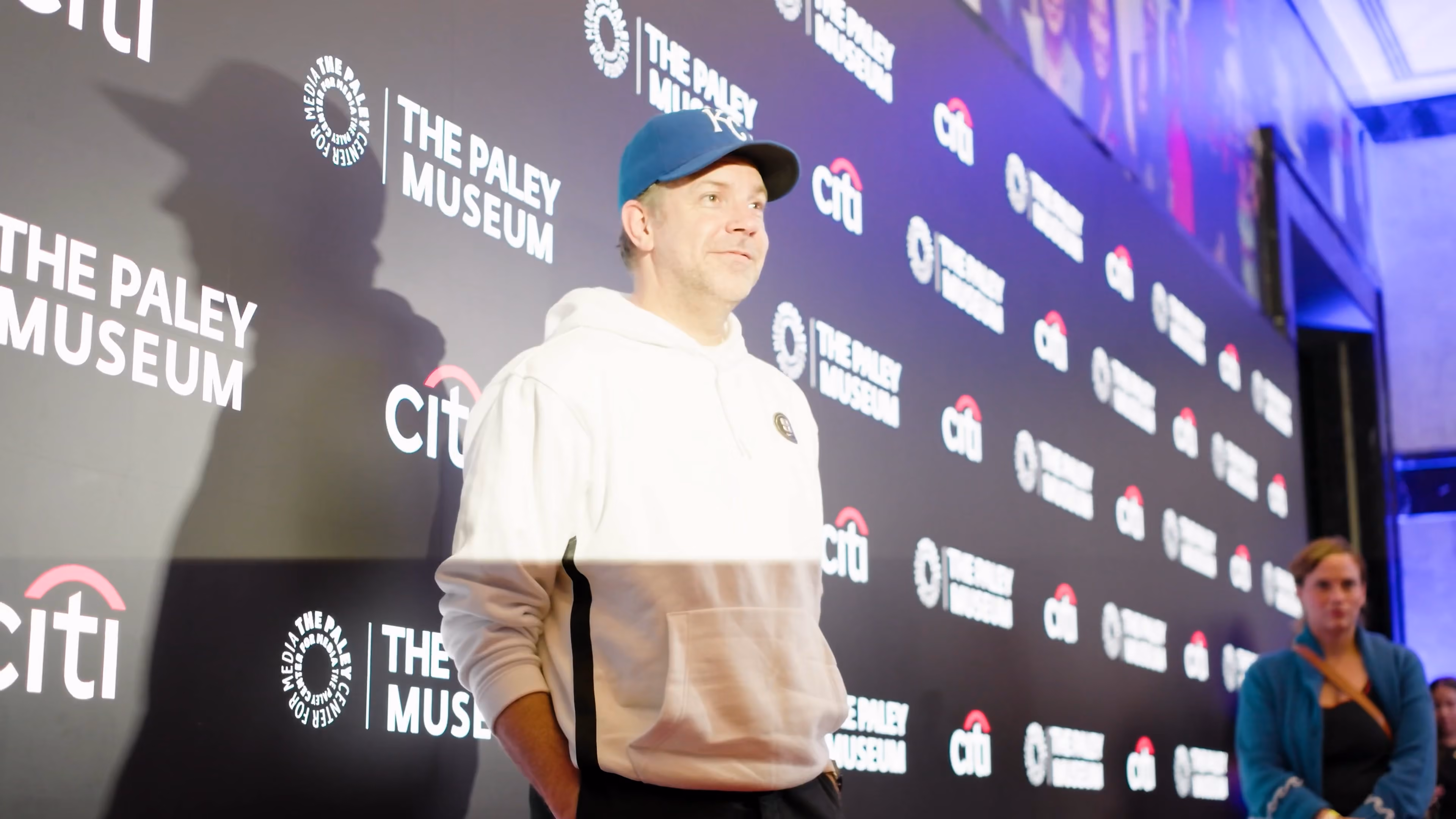 Jason Sudeikis in a white hoodie and blue cap stands by a wall with The Paley Museum and Citi logos.