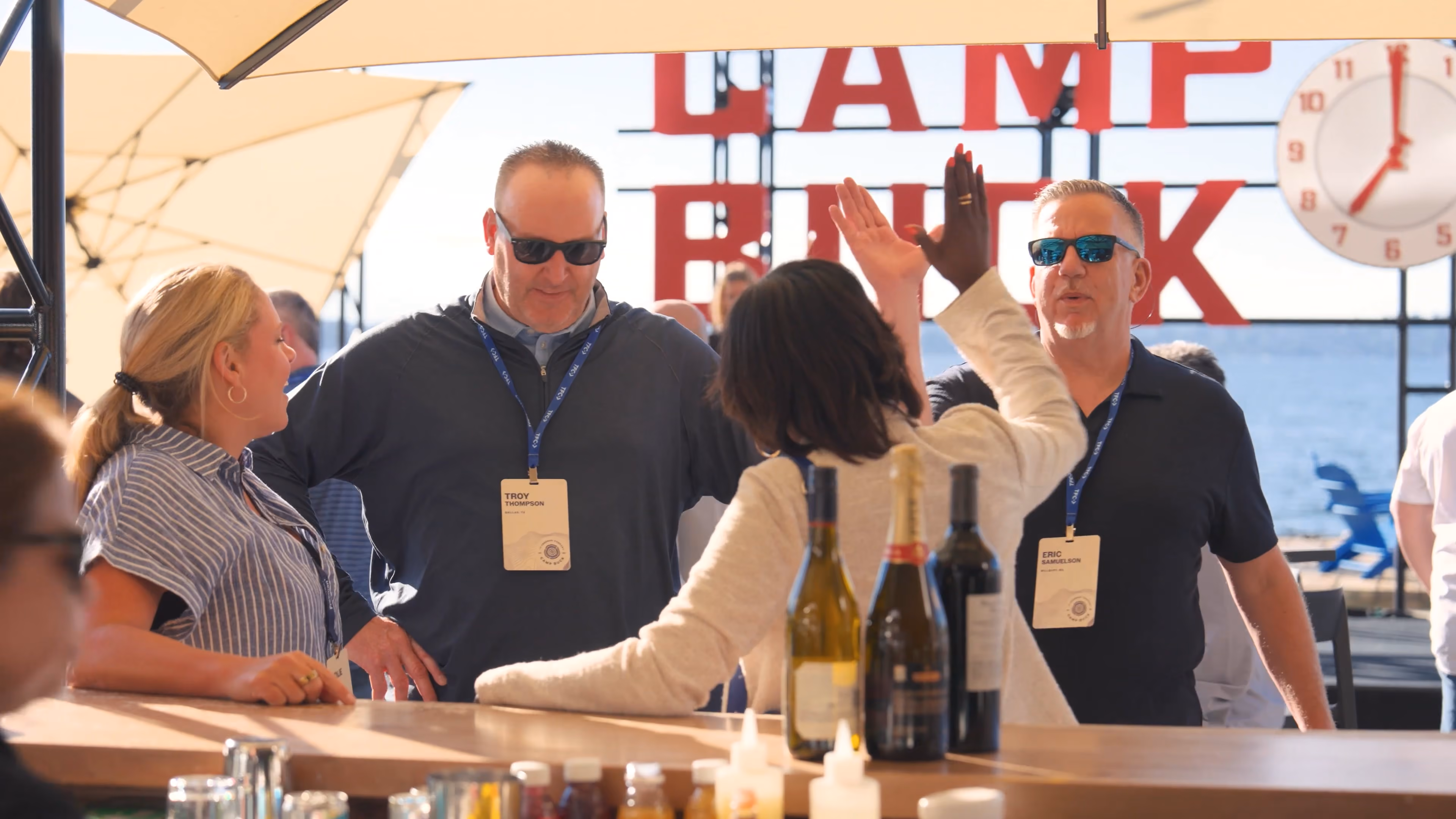 People socializing at an outdoor bar with drinks and umbrellas on a sunny day near water.