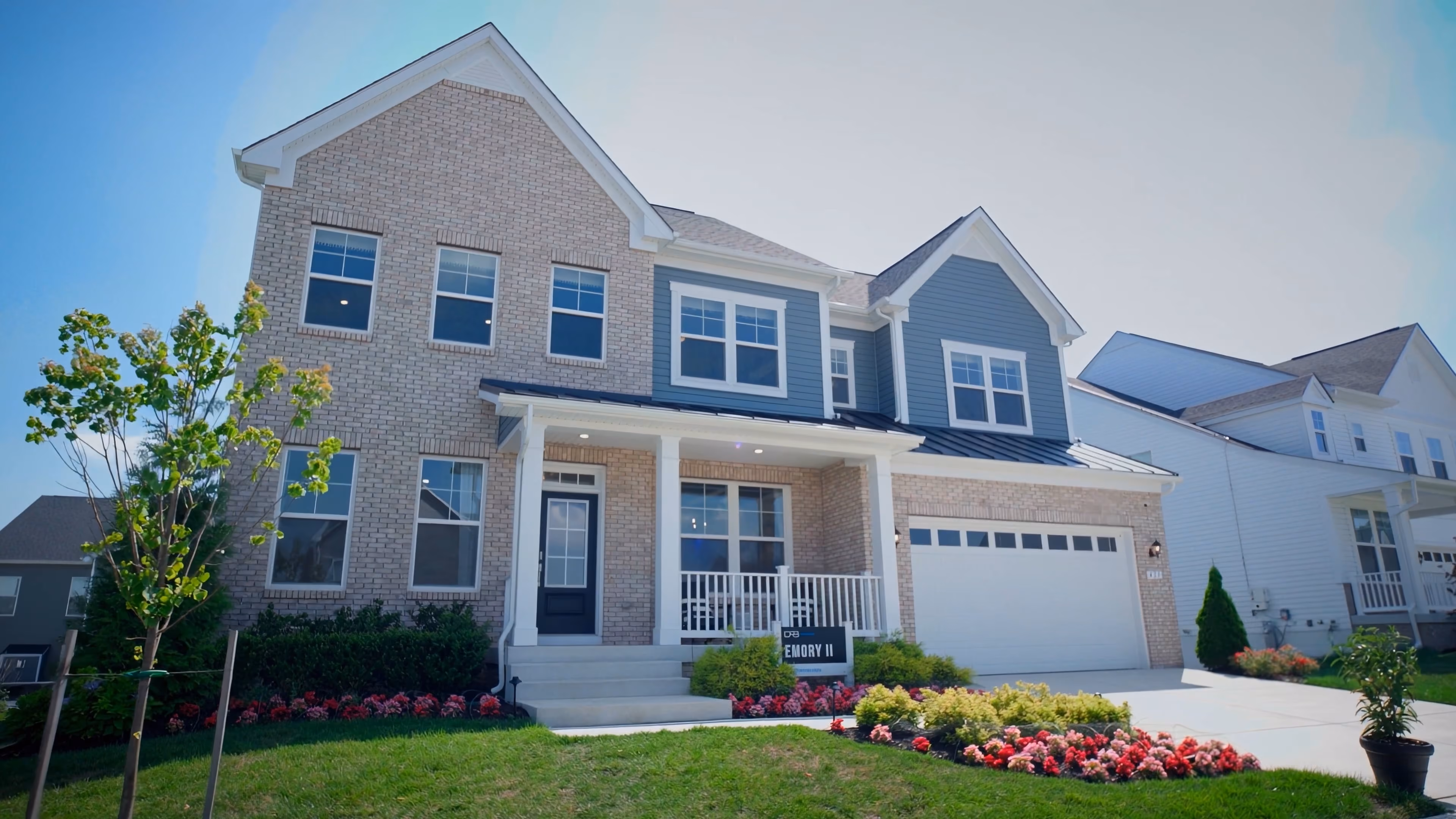 Modern two-story house with brick and blue siding, front porch, garage, and landscaped yard with flowers.