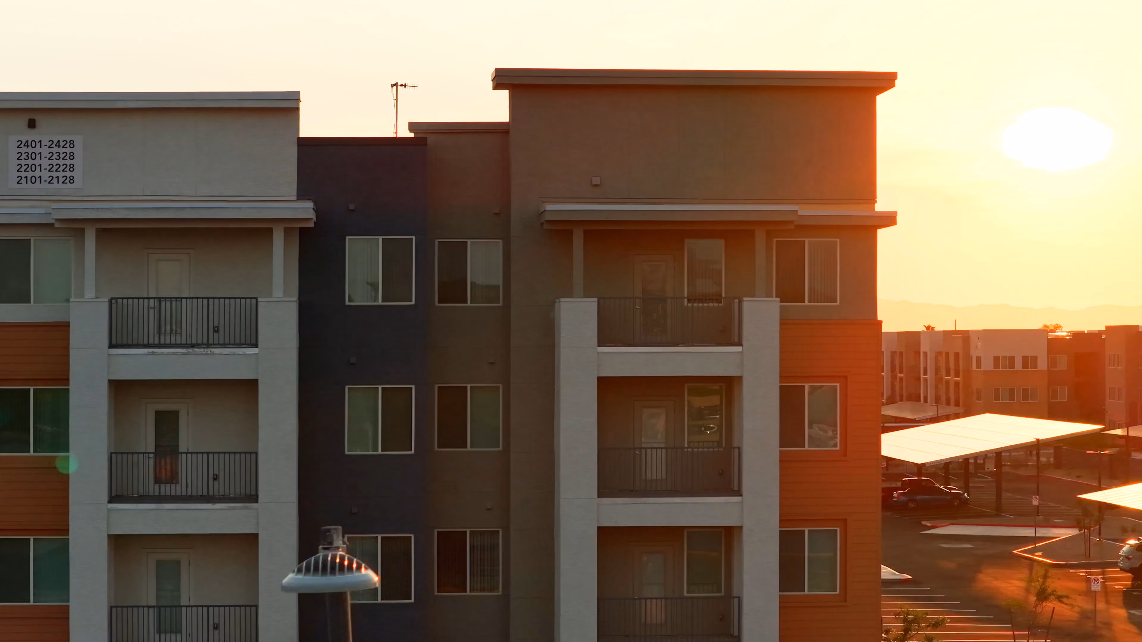 Modern three-story apartment building in warm sunset light with balconies, parking lot, and nearby buildings.
