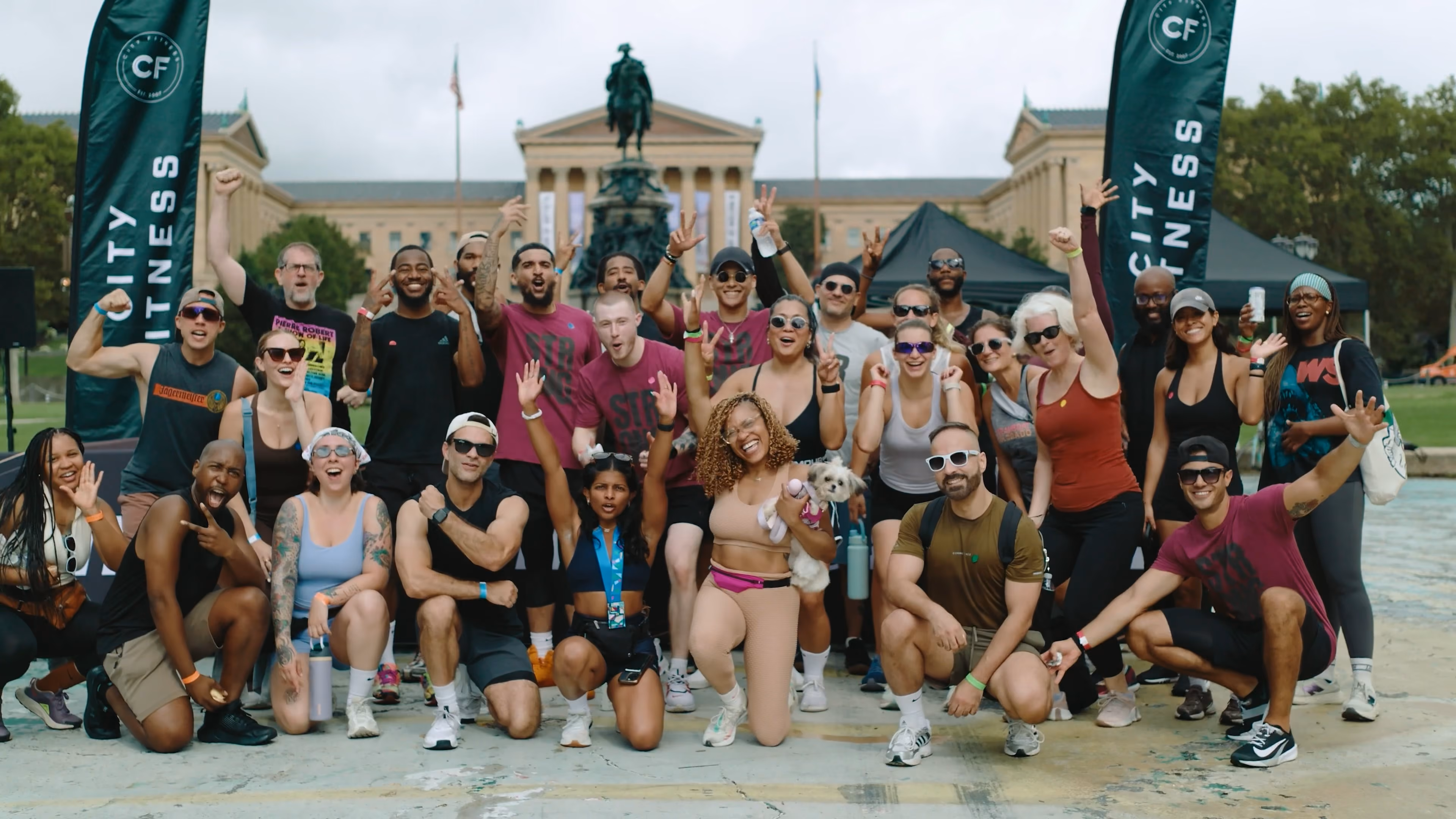 Group of diverse men and women posing with smiles and raised hands outdoors in front of a classical building with a statue, two vertical banners reading 'CITY FITNESS' are visible on each side.