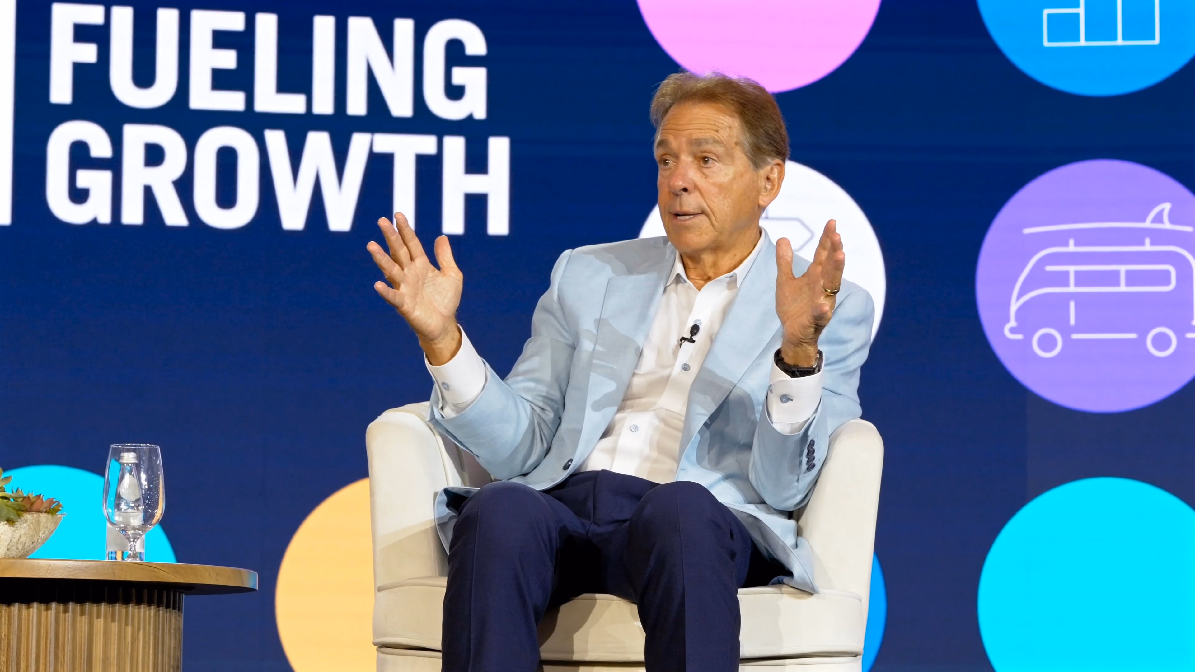 A man in a light blue blazer and dark trousers sits on a white armchair with his hands raised, speaking at an event with a backdrop displaying the words 'FUELING GROWTH' and colorful icons including a bus.