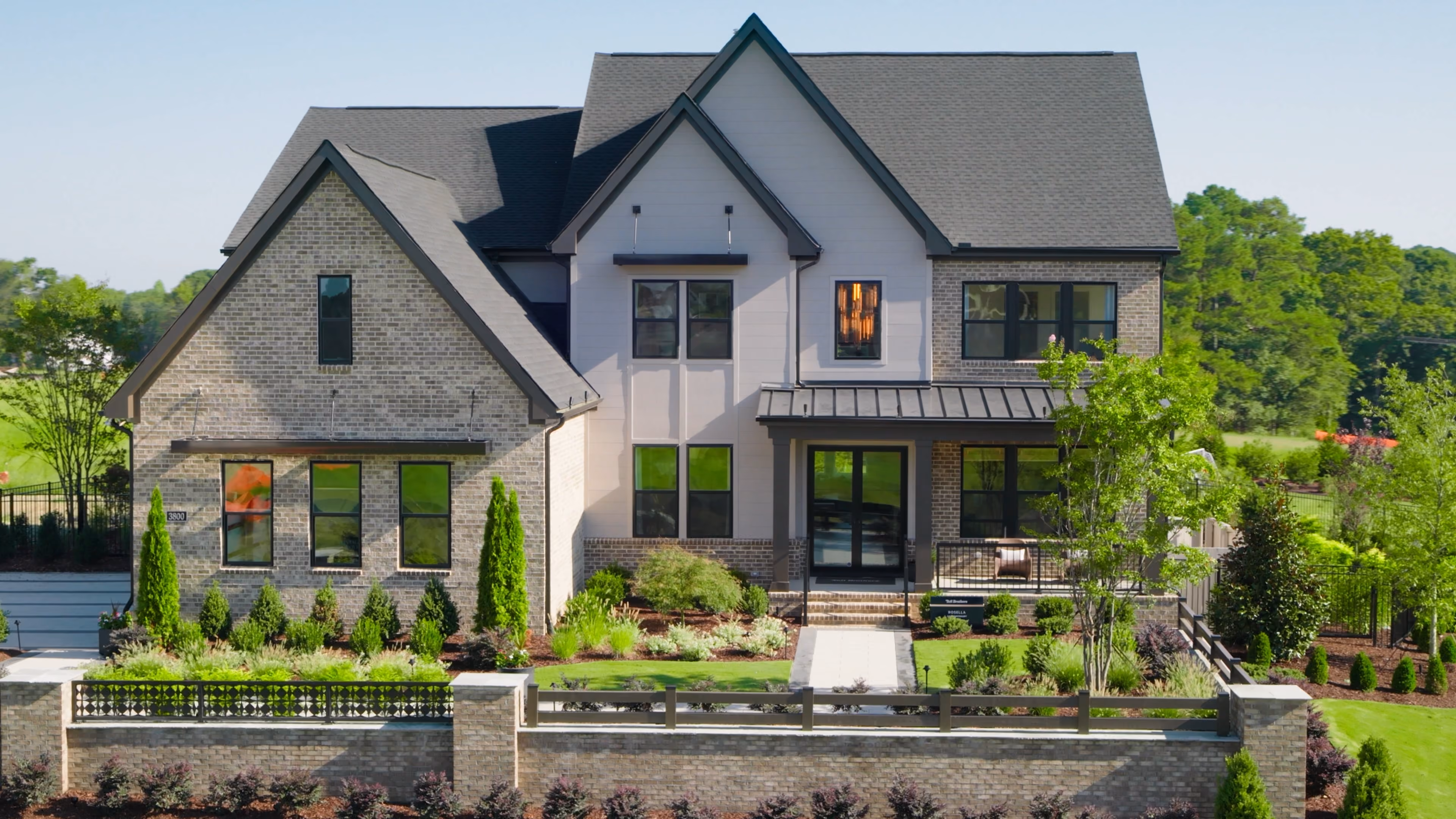 Modern two-story house with a combination of light brick and white siding exterior, a dark gray roof, and multiple windows, surrounded by a landscaped yard with green grass, shrubs, small trees, and a stone fence at the front.