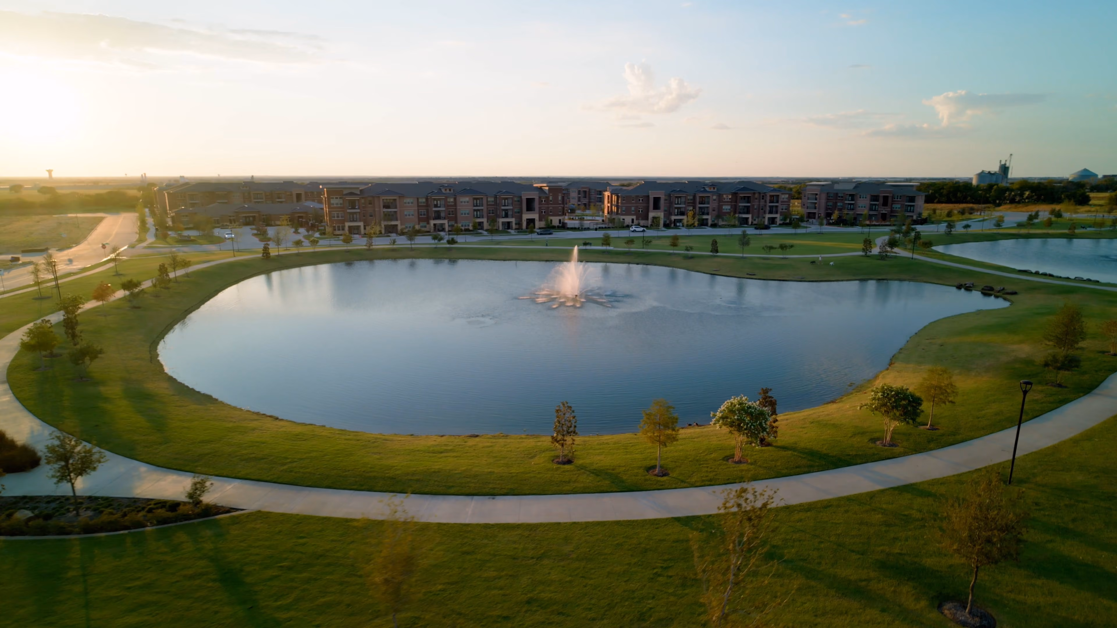 Aerial view of a circular pond with a central water fountain, surrounded by a walking path and green grass, with modern apartment buildings in the background under a clear sky at sunset.