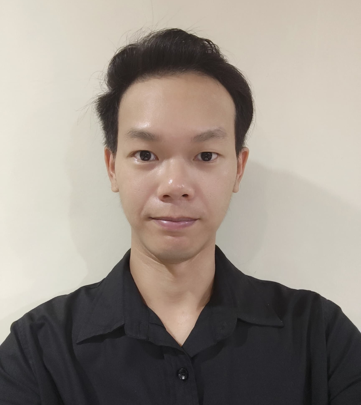 Young man with short black hair wearing a black collared shirt, posing against a plain beige wall.