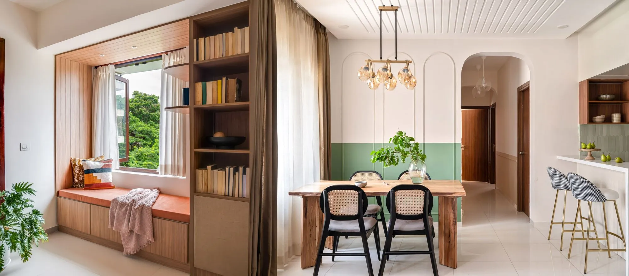 Modern dining area with wooden table, four black chairs, a green two-tone wall, pendant lights, and a cozy window seat with cushions and a throw blanket.